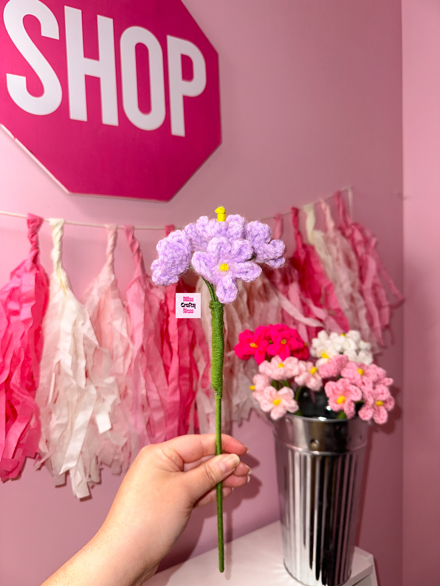 Hand holding crocheted flower with a pink 'SHOP' sign in the background.
