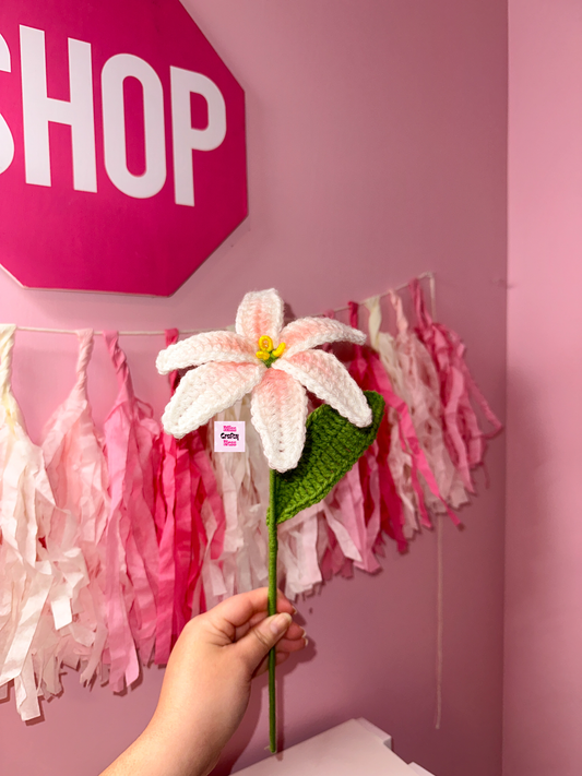 Hand holding a crochet lily flower against a pink wall with 'SHOP' sign and tassels.