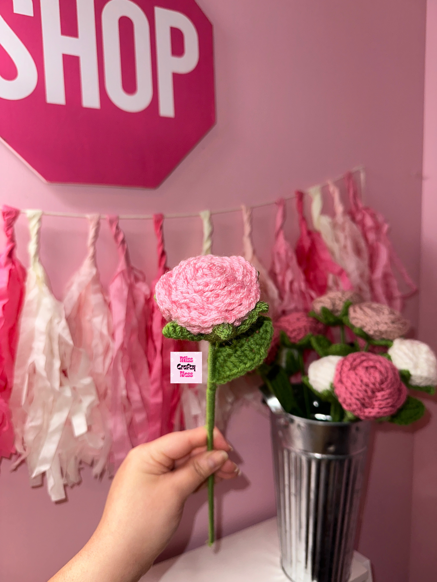 Hand holding a pink crochet rose flower with a pink 'SHOP' sign and floral decorations in the background.