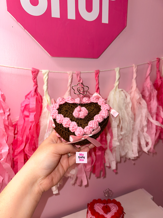 Brown Heart-shaped crochet cake with pink frosting held in front of a pink wall with tassels.