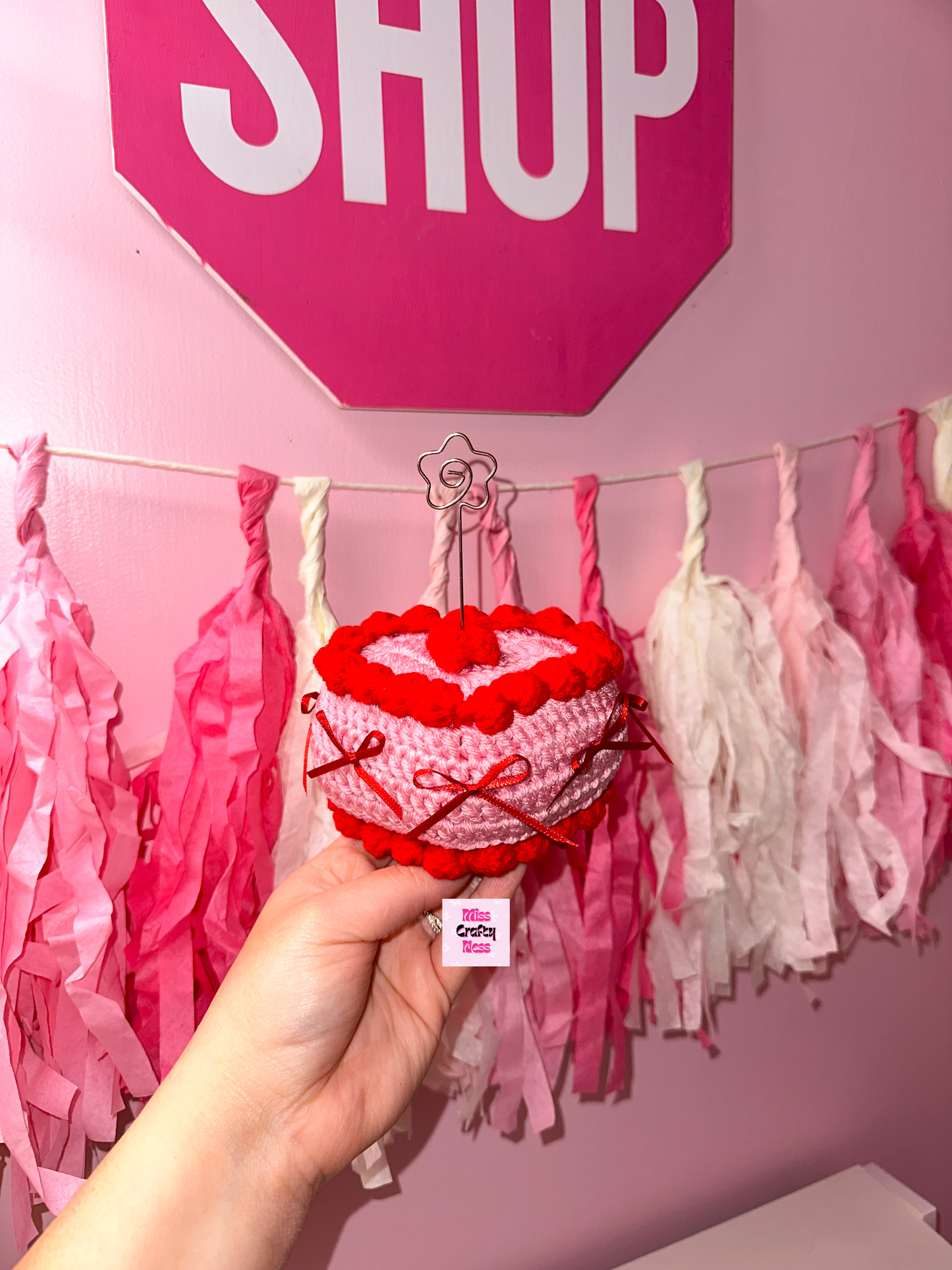Heart-shaped crochet cake held by a hand with pink and white tassel decorations on a pink wall.