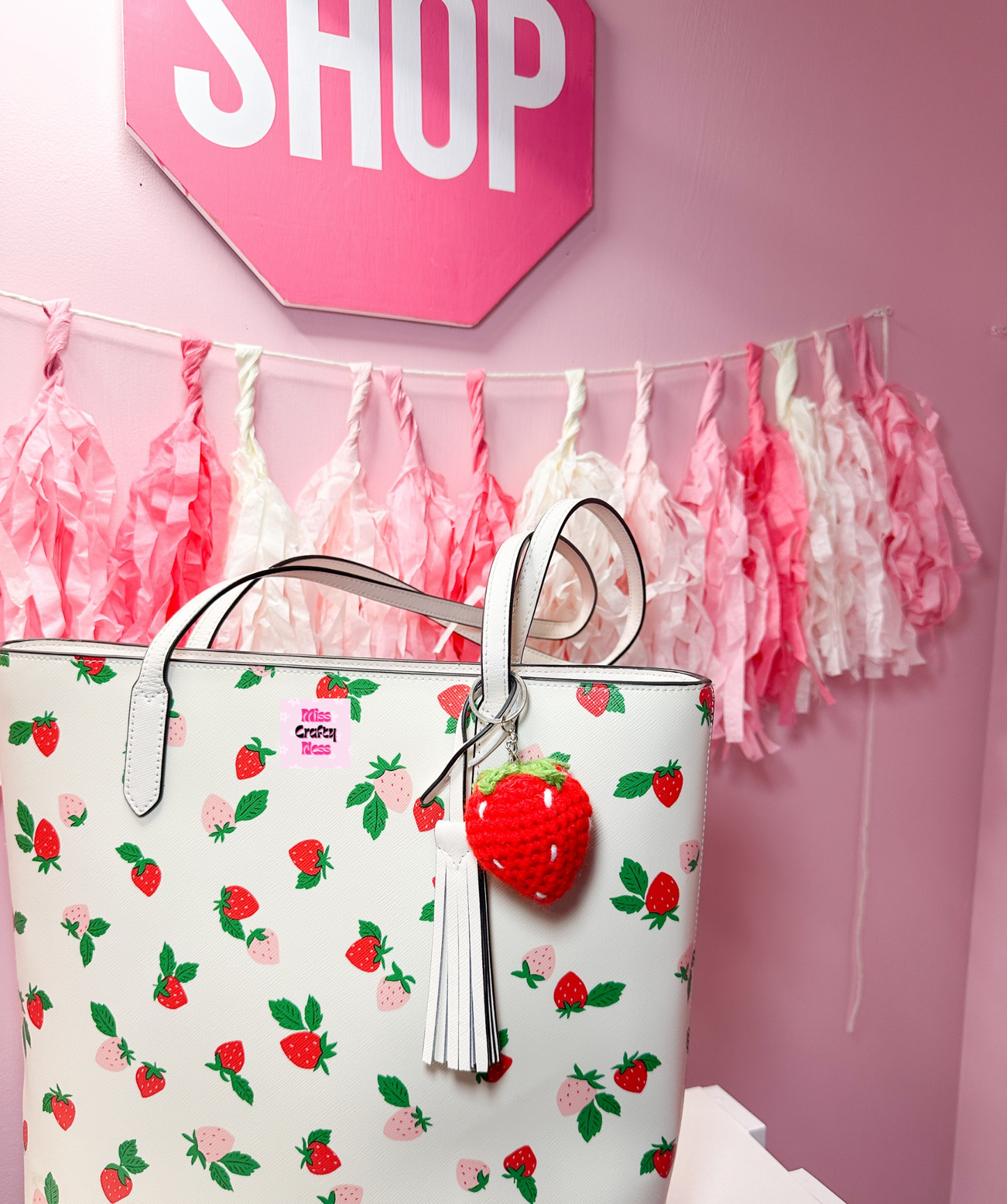 White tote bag with strawberry pattern and red strawberry keychain against a pink wall with 'SHOP' sign and tassels.