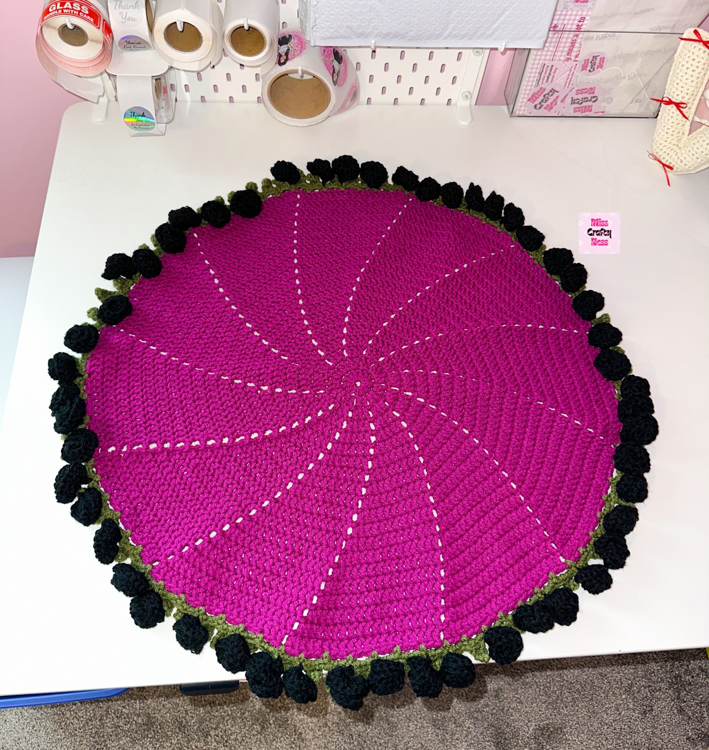 Round violet crochet blanket with black roses and green leaves on a table with shipping supplies.
