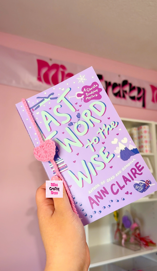 Hand holding a purple book with a heart shaped string bookmark wrapped around it, with a decorative background.