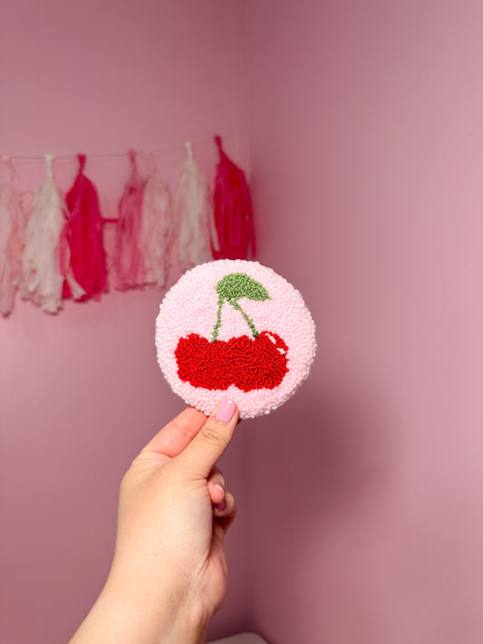 Hand holding a Round cherry coaster, with a pink background and decorative tassels.