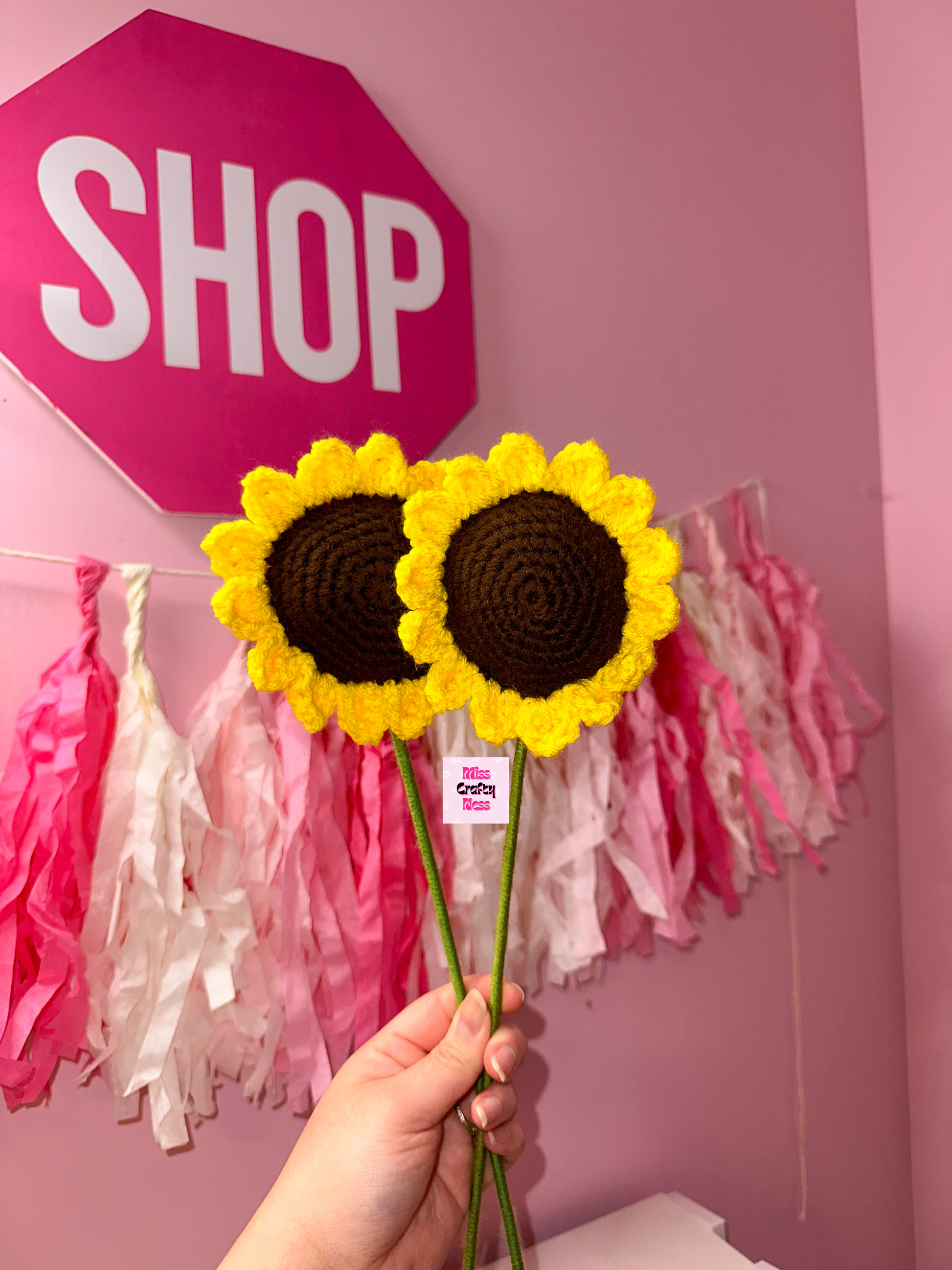 Hand holding crochet sunflowers with a pink wall and 'SHOP' sign in the background