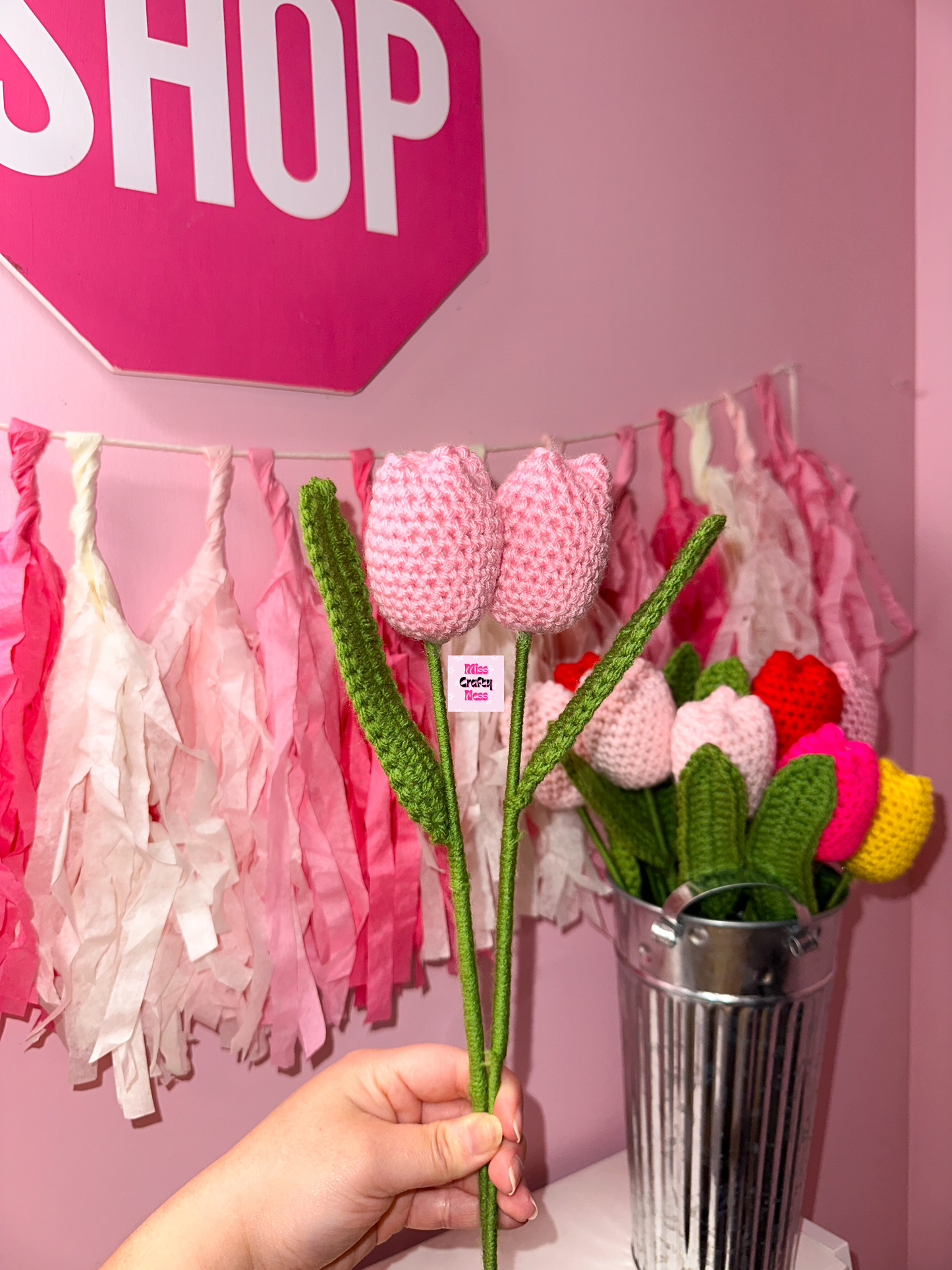 Hand holding a crocheted tulip flower with a pink 'SHOP' sign and decorative tassels in the background.