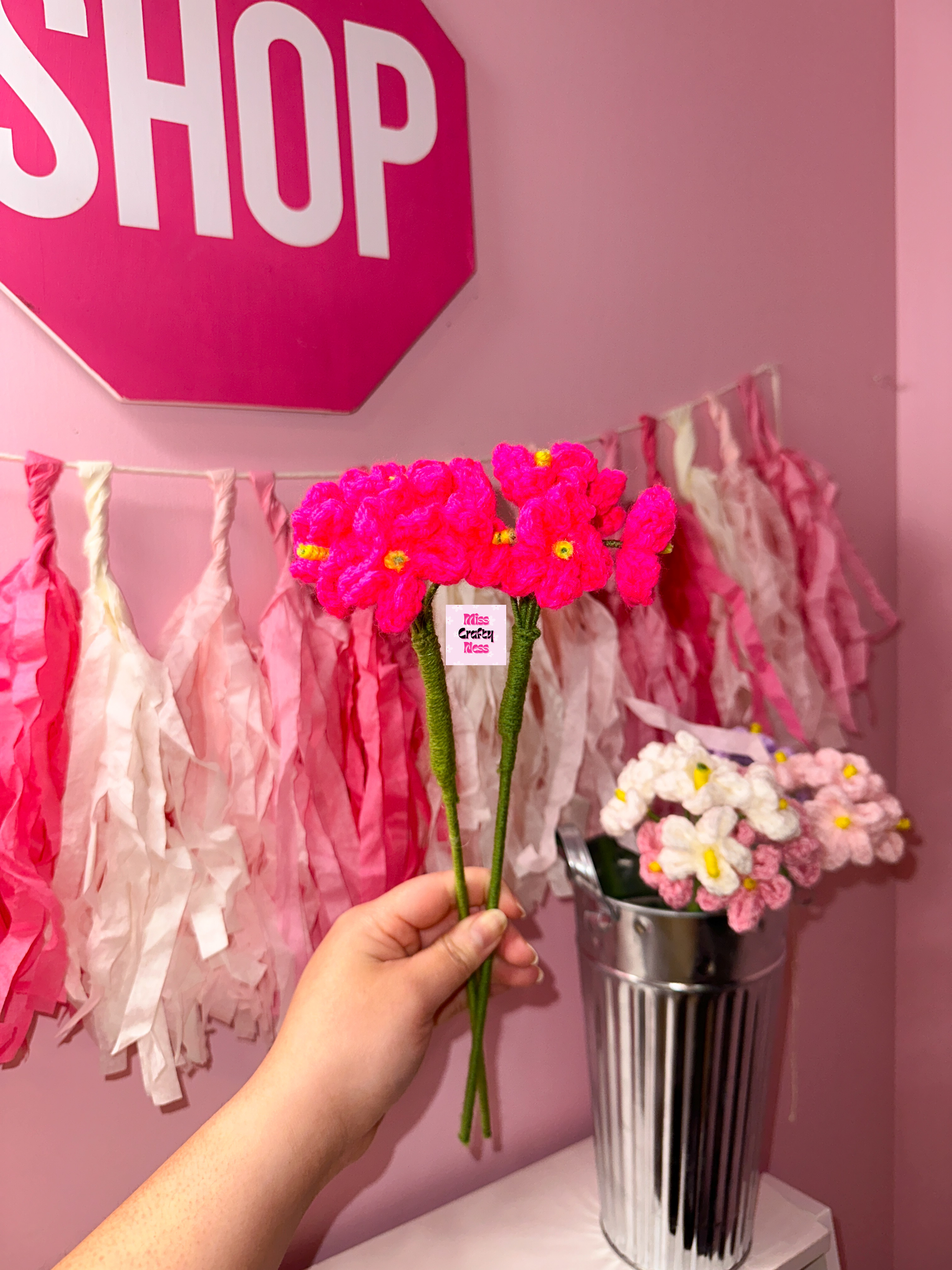 Hand holding pink crochet flowers in front of a pink wall with 'SHOP' sign and decorative tassels.