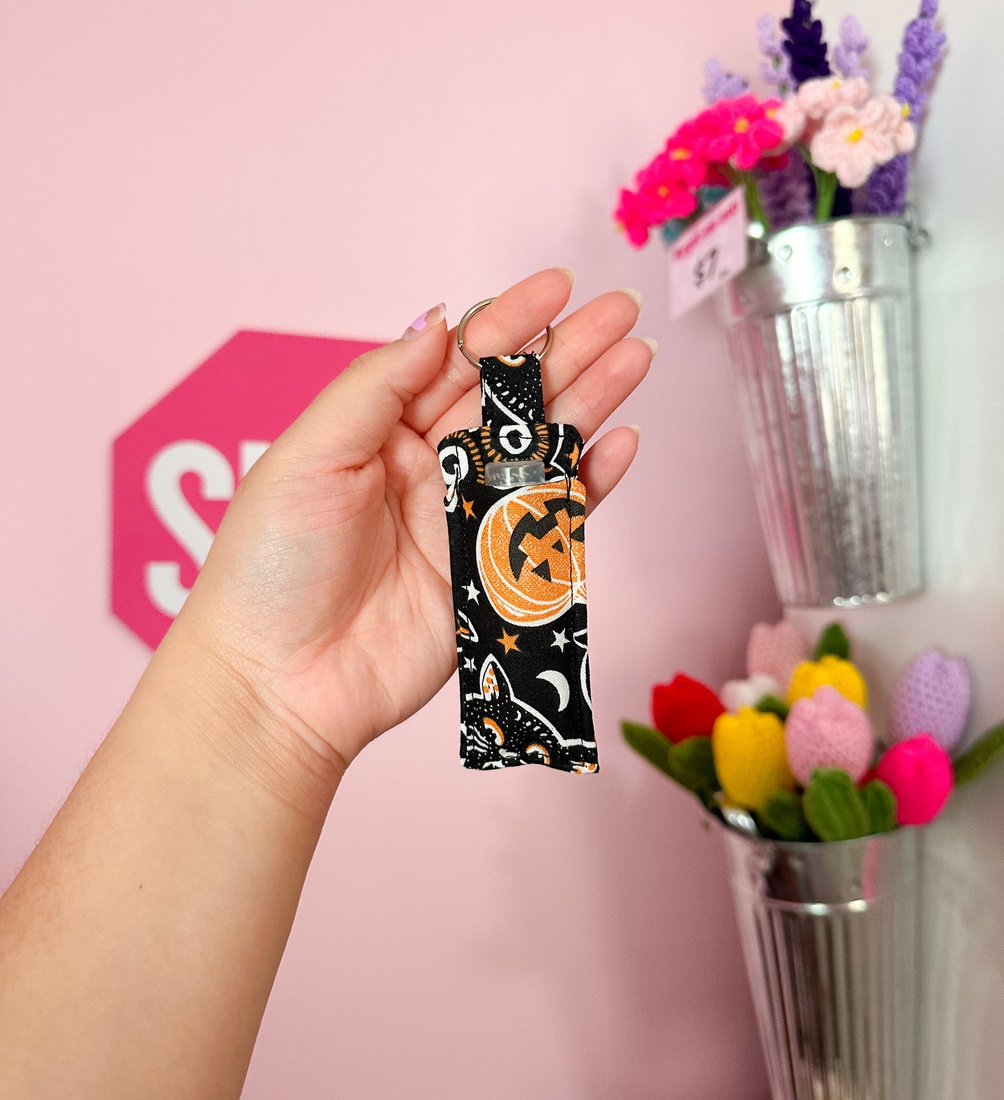 Hand holding a lip balm holder keychain, keychain has a Halloween themed pattern with a pink 'SHOP' sign and tassels in the background
