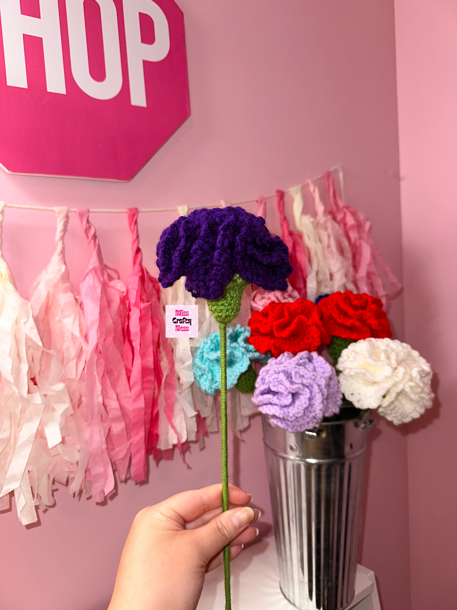 Hand holding a dark purple crocheted flower in front of a pink wall with tassels.