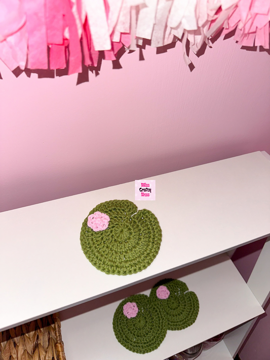 Three green crocheted coasters with pink flowers on a white surface, with a pink tassels in the background.