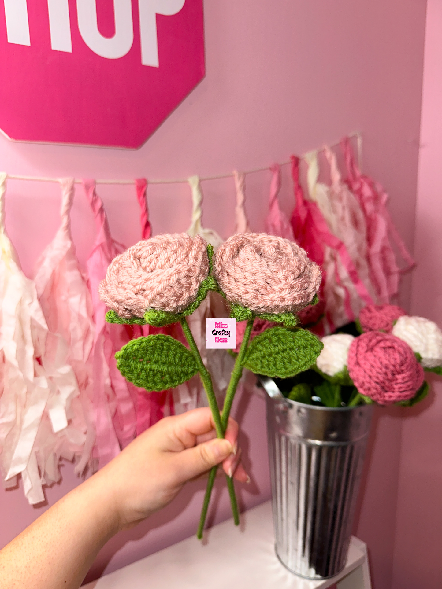 Hand holding a pink crochet rose flower with a pink 'SHOP' sign and floral decorations in the background.