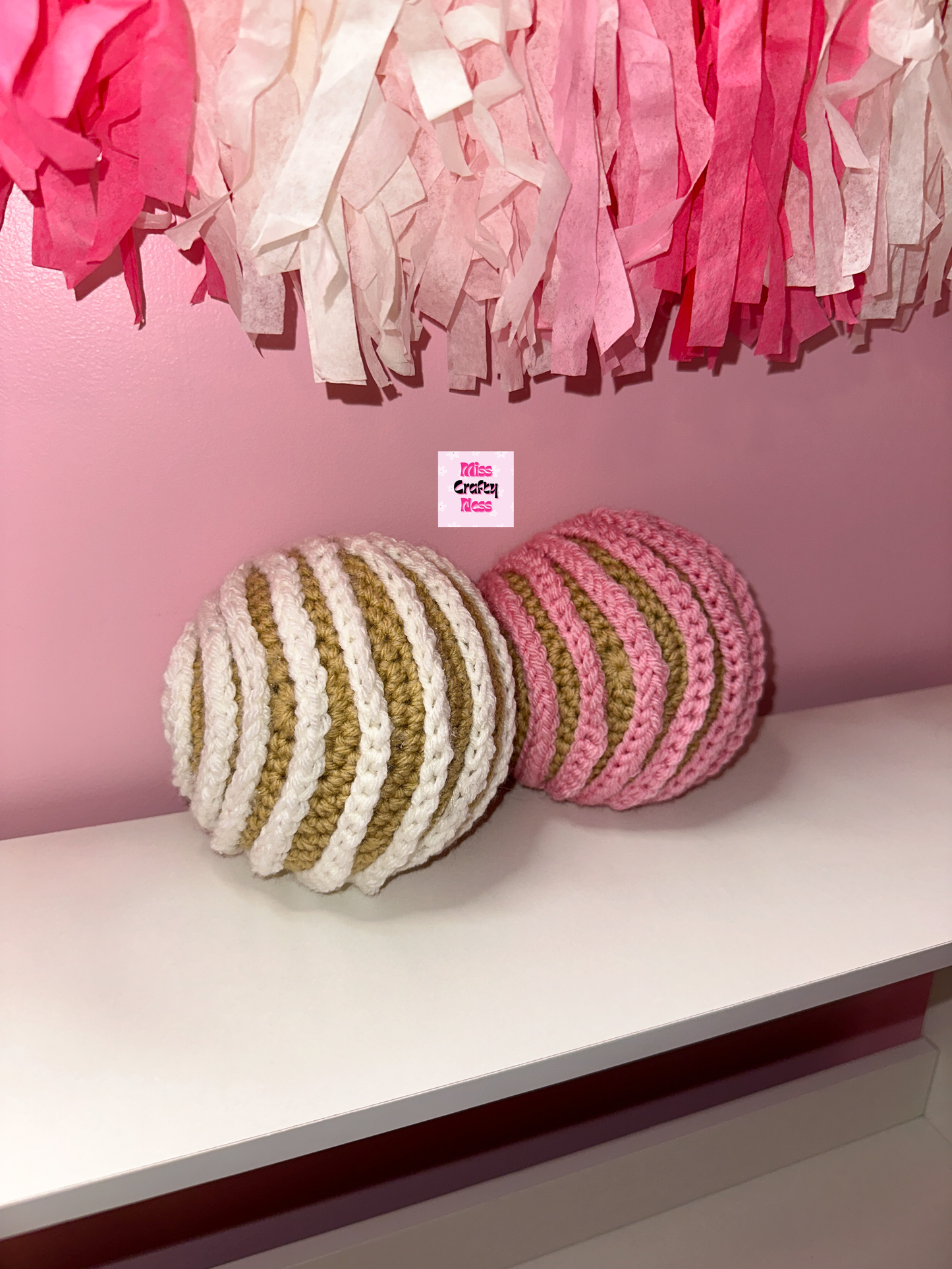 Two crocheted sweet breads one pink and one white, on a shelf against a pink wall with tassels.