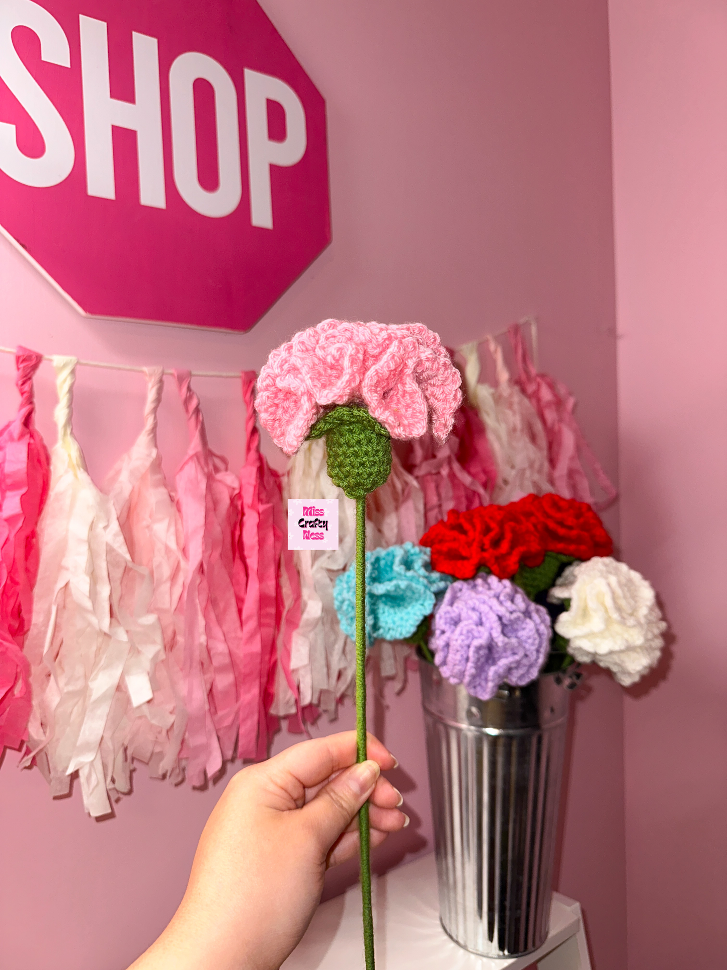 Hand holding a crochet flower in front of a pink wall with 'SHOP' sign and decorative items.