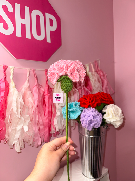 Hand holding a crochet flower in front of a pink wall with 'SHOP' sign and decorative items.