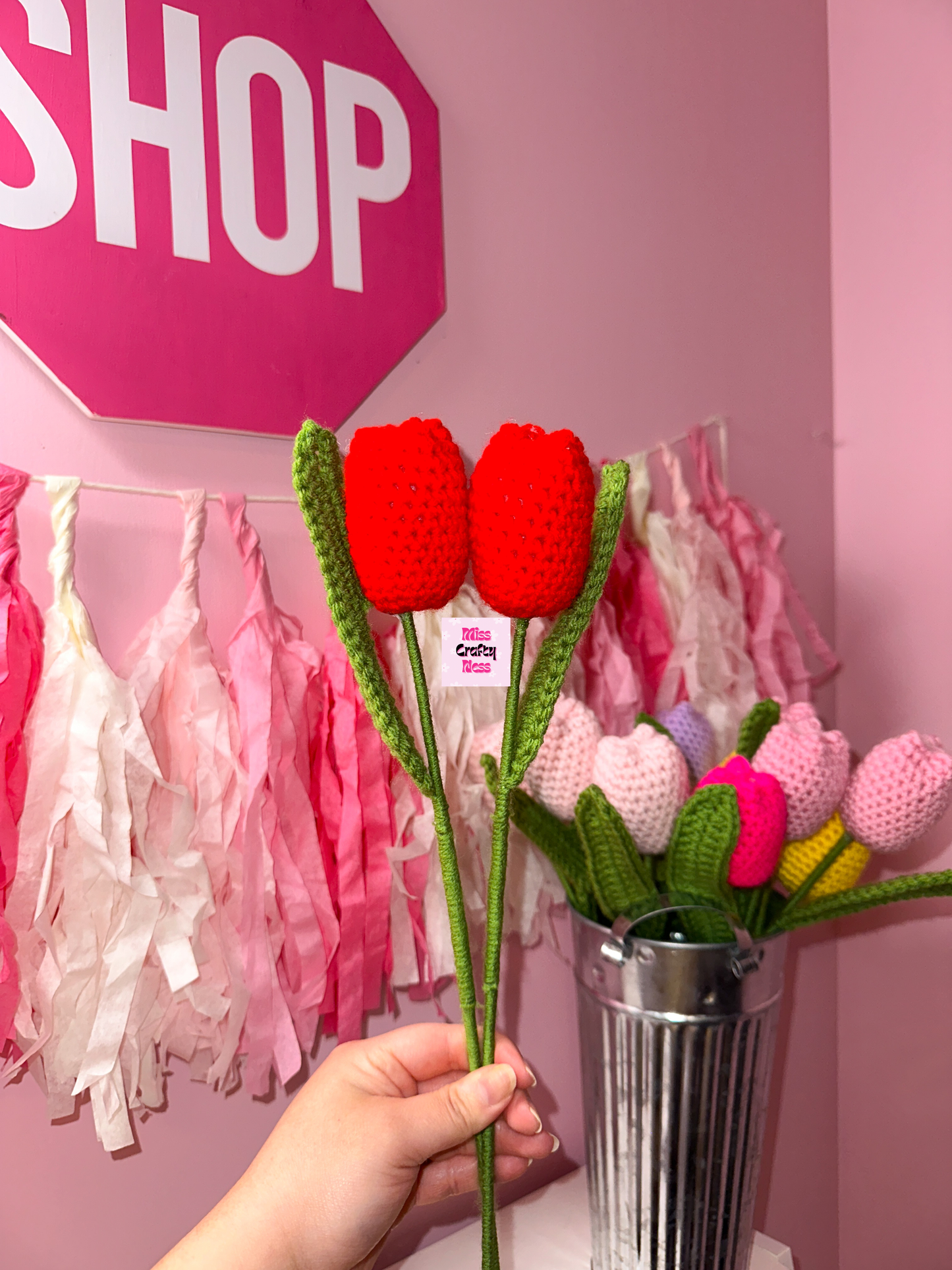 Hand holding crocheted tulip flowers with a pink 'SHOP' sign and decorative tassels in the background.