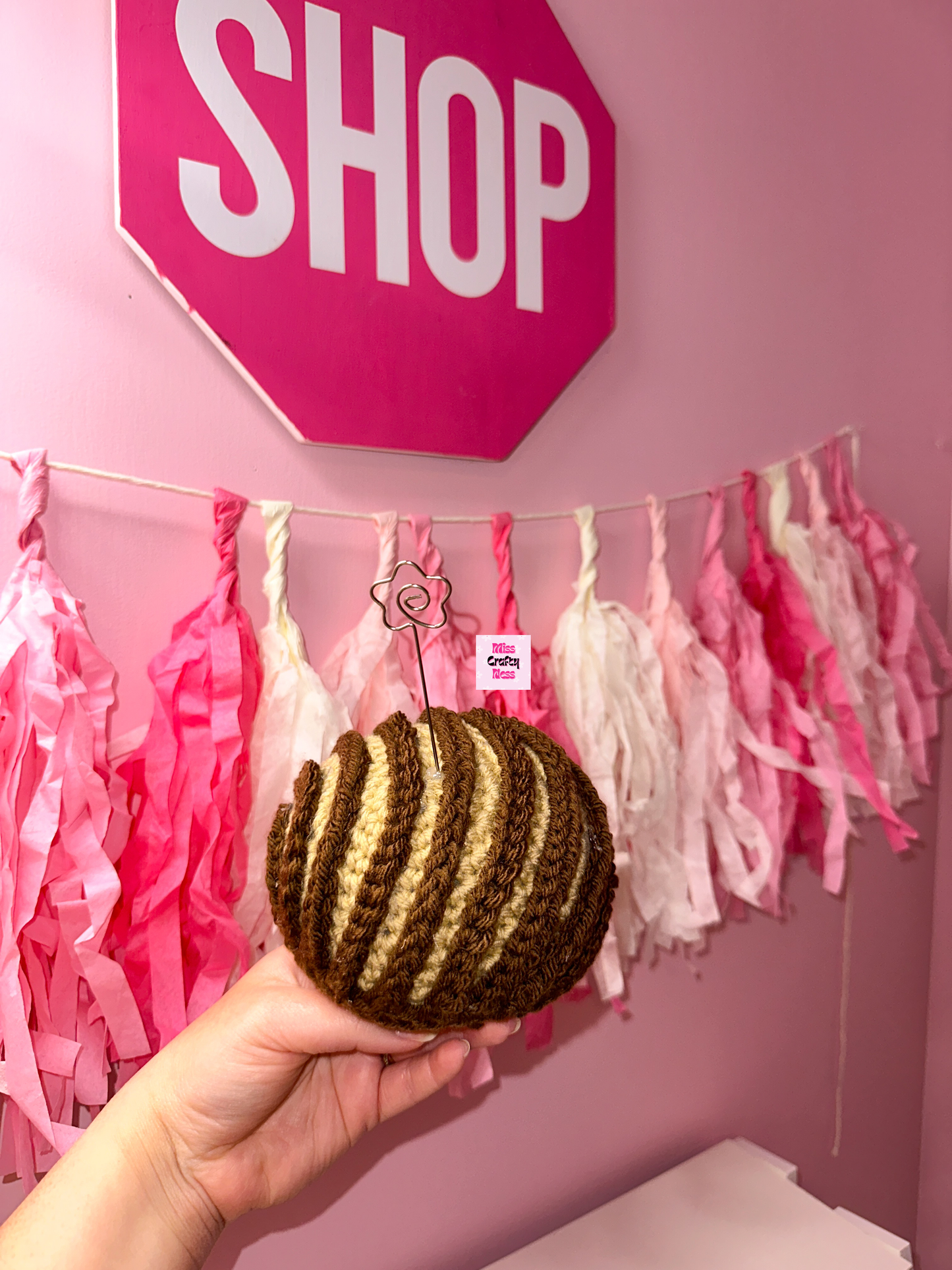 Crochet sweet bread photo holder item with pink and white tassels and a sign that reads ‘SHOP’ in the background.