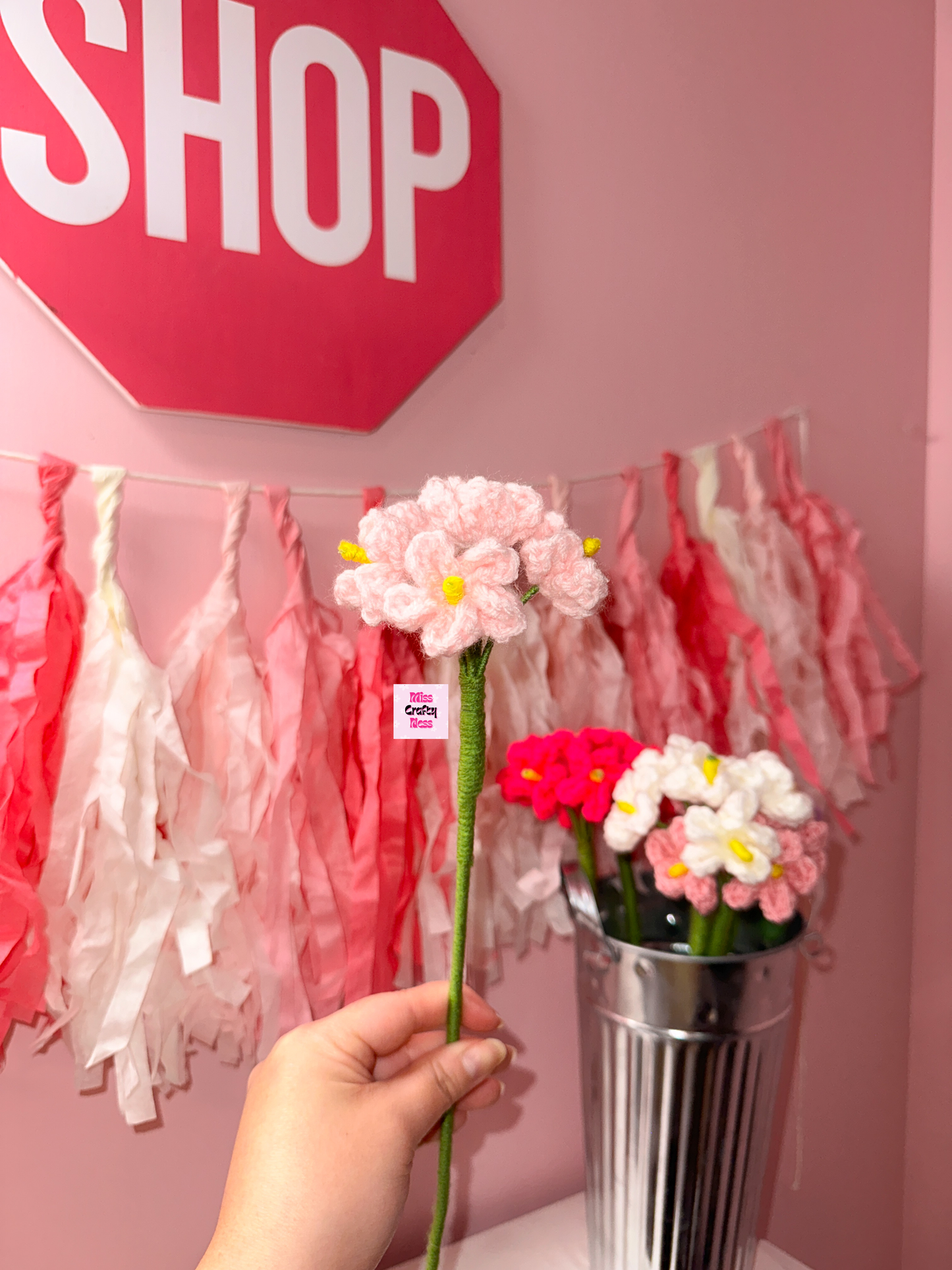 Hand holding crocheted flower with a pink 'SHOP' sign in the background.