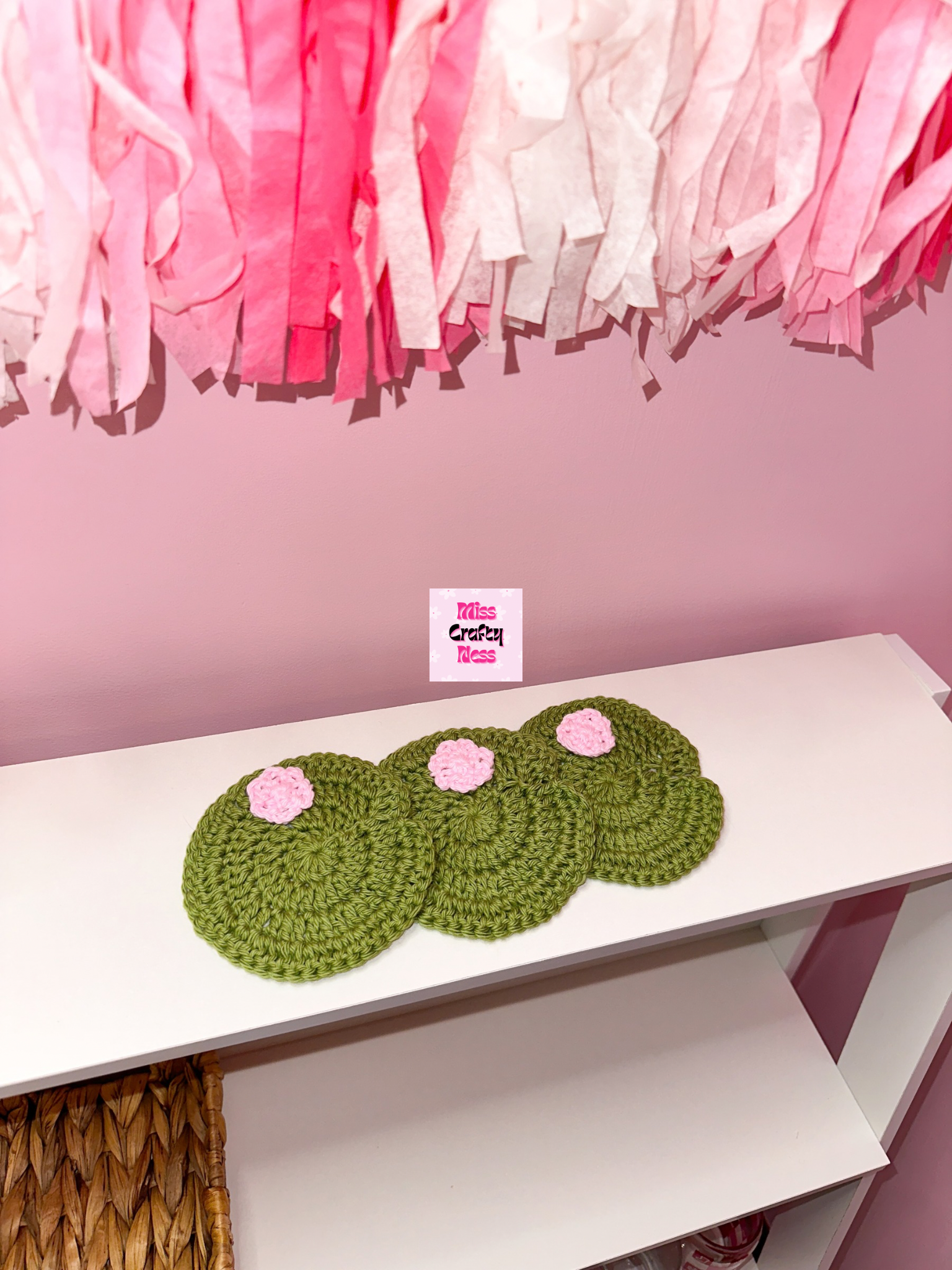 Three green crocheted coasters with pink flowers on a white surface, with a pink tassels in the background.