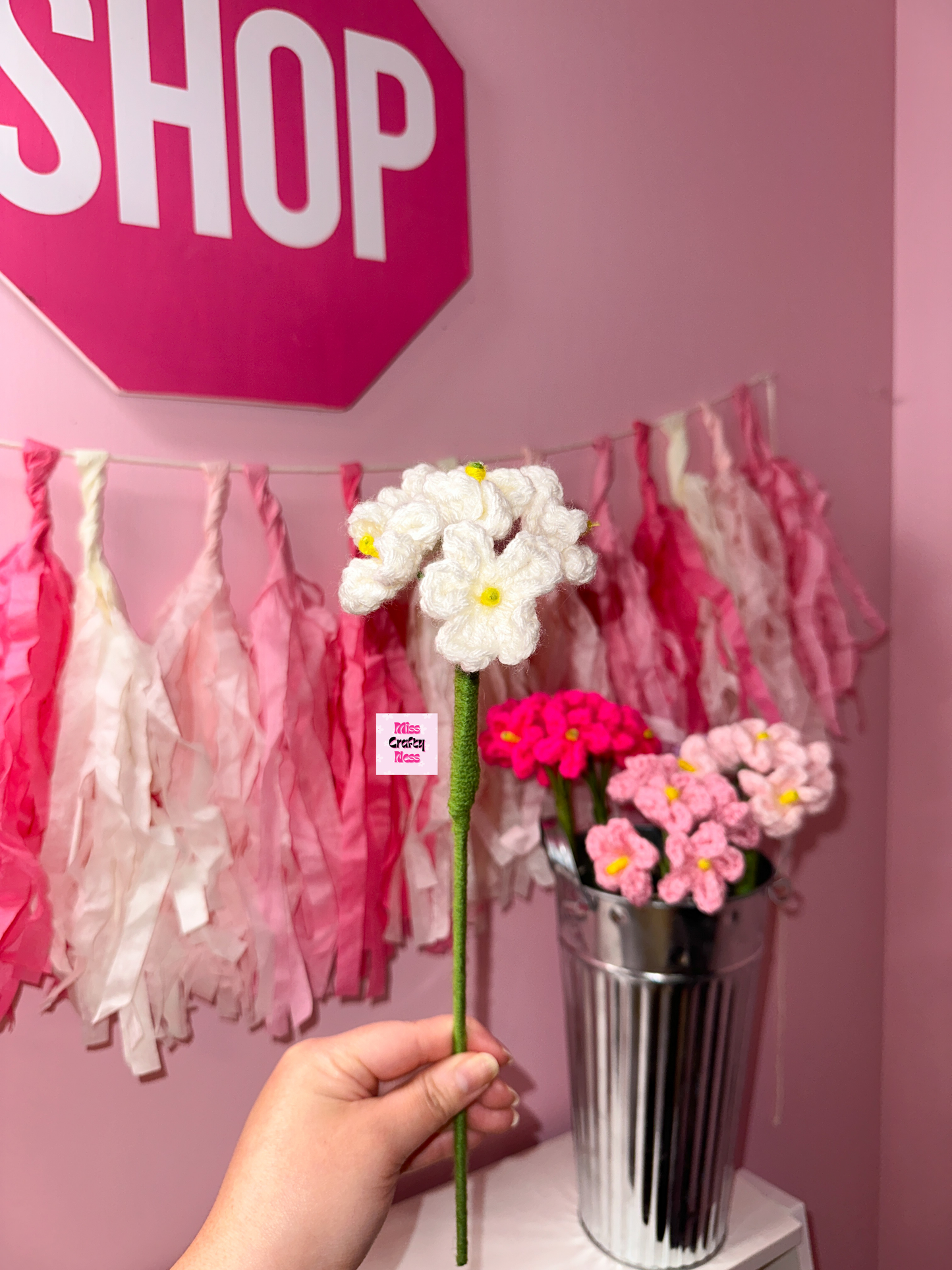 Hand holding crocheted flower with a pink 'SHOP' sign in the background.