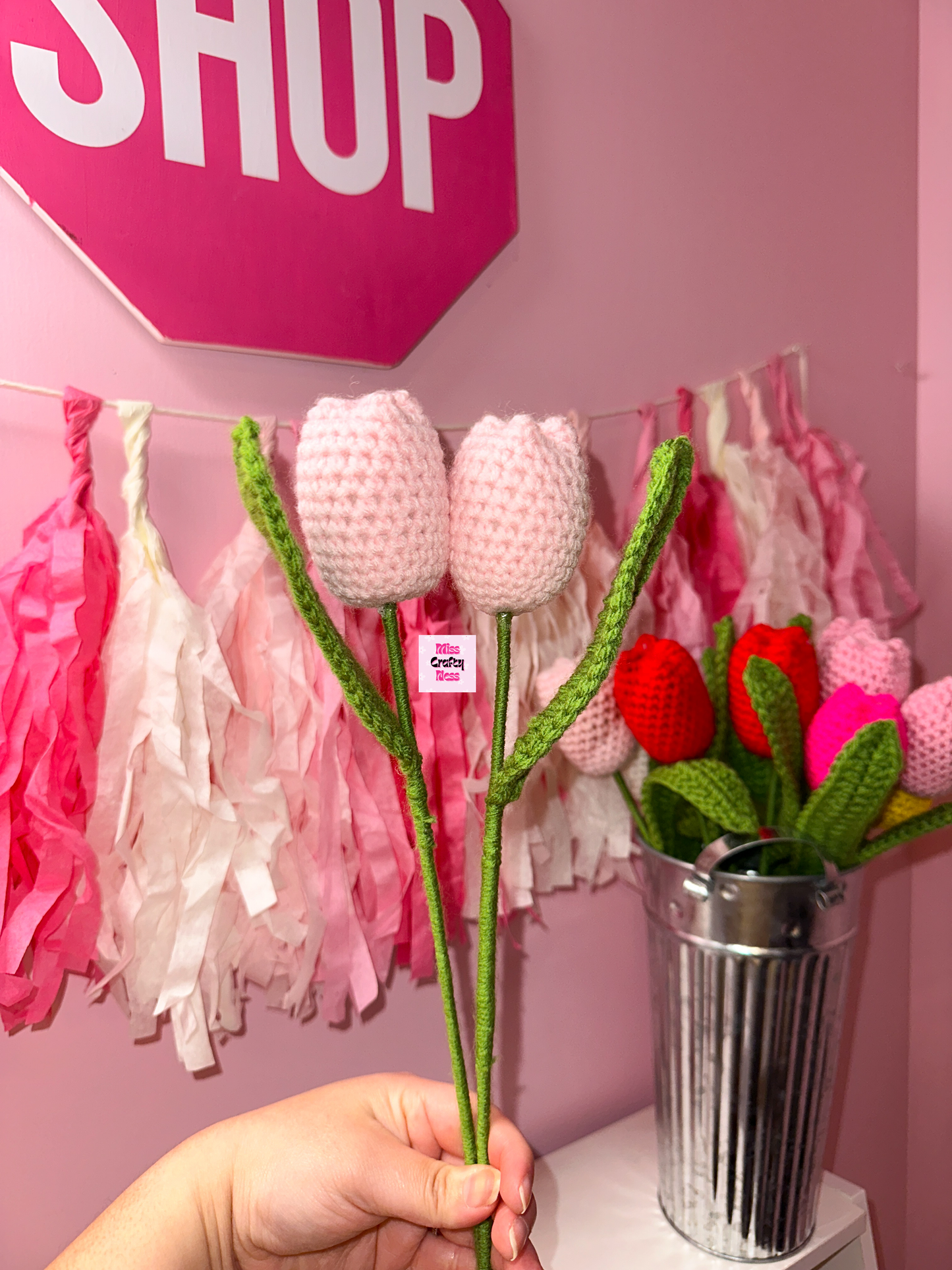 Hand holding crocheted tulip flowers with a pink 'SHOP' sign and decorative tassels in the background.