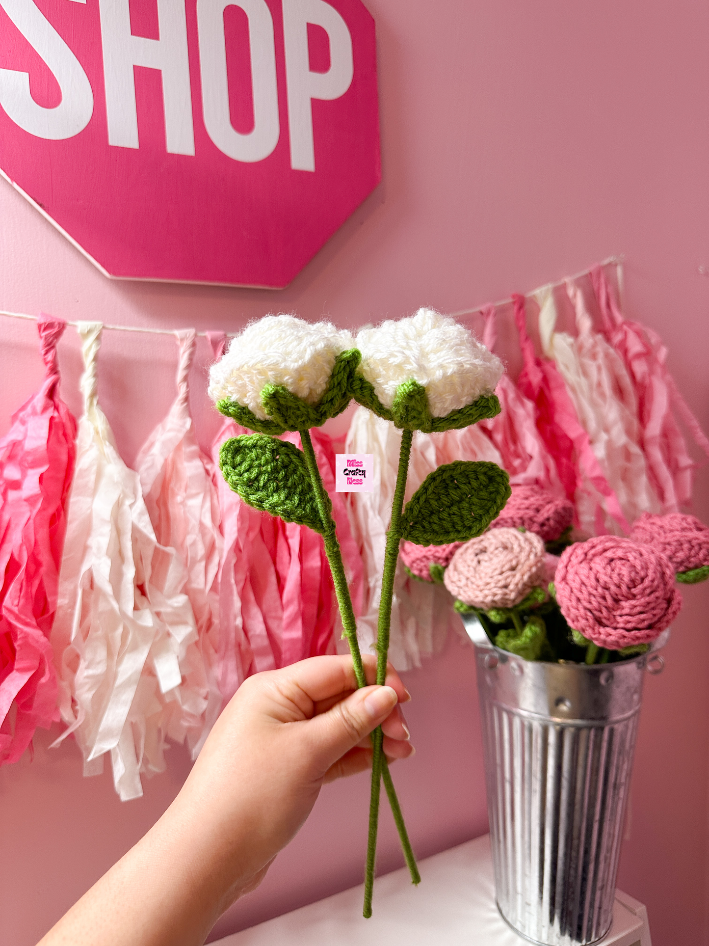 Hand holding a crocheted white rose flower with a pink 'SHOP' sign and decorative tassels in the background.