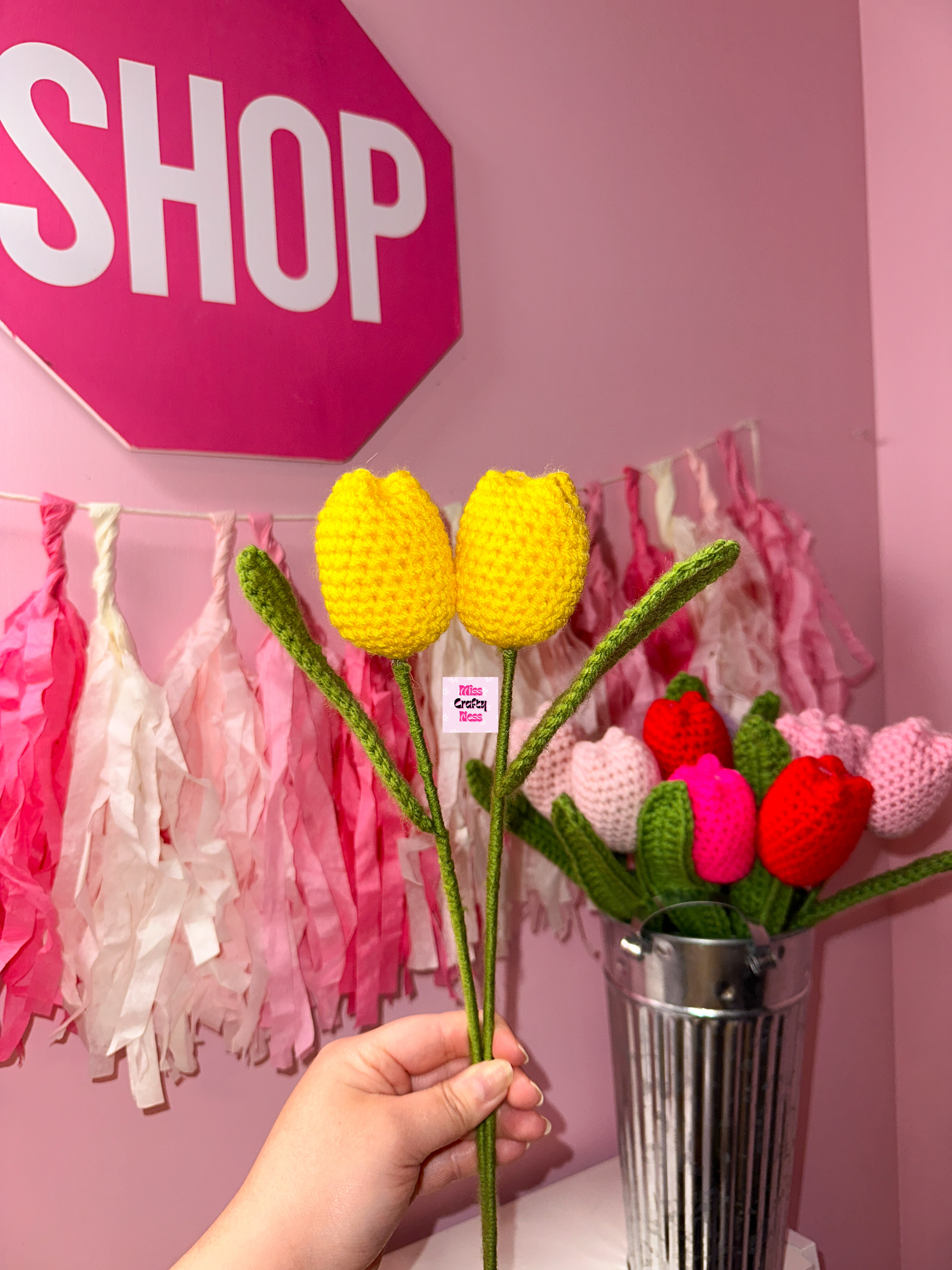Hand holding crocheted tulip flowers with a pink 'SHOP' sign and decorative tassels in the background.