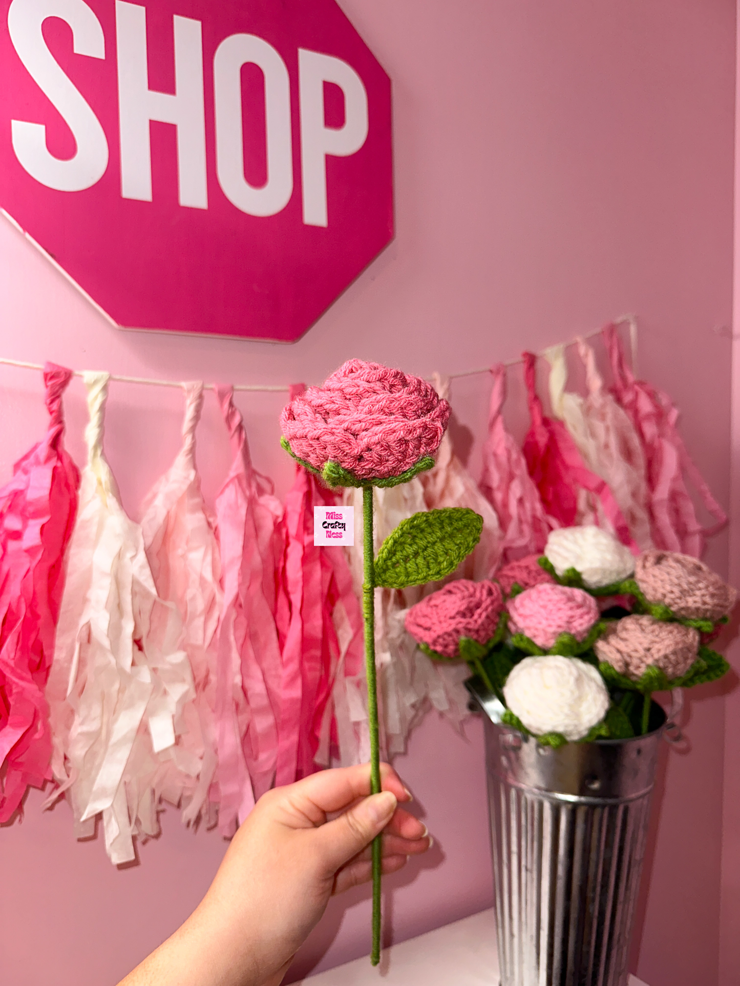 Hand holding a dark pink crochet rose flower with a pink 'SHOP' sign and floral decorations in the background.