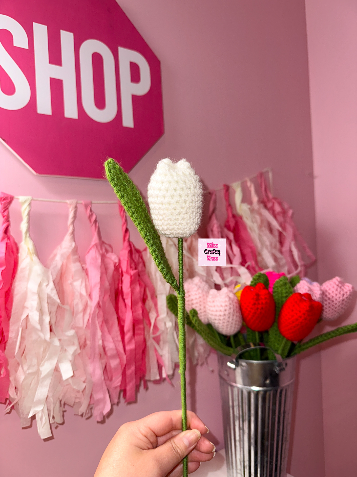 Hand holding a crocheted tulip flower with a pink 'SHOP' sign and decorative tassels in the background.