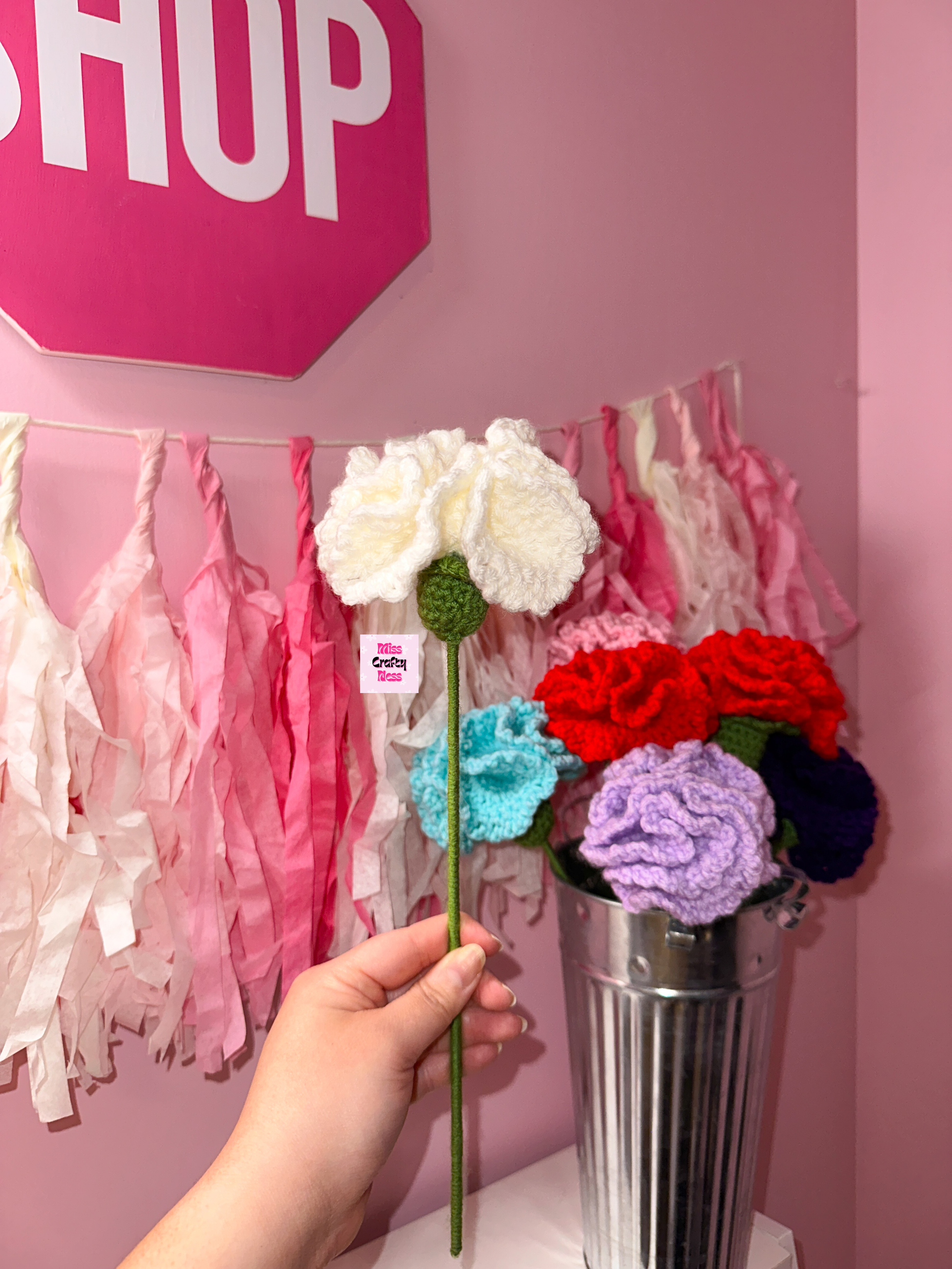 Hand holding a white crocheted flower in front of a pink wall with tassels.