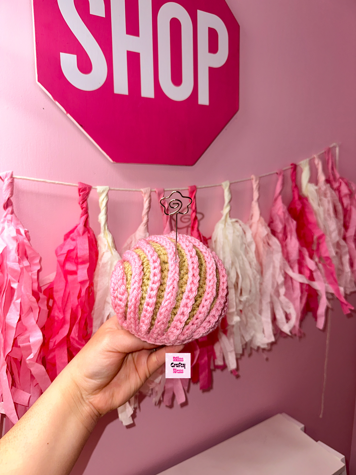 Crochet sweet bread photo holder item with pink and white tassels and a sign that reads ‘SHOP’ in the background.