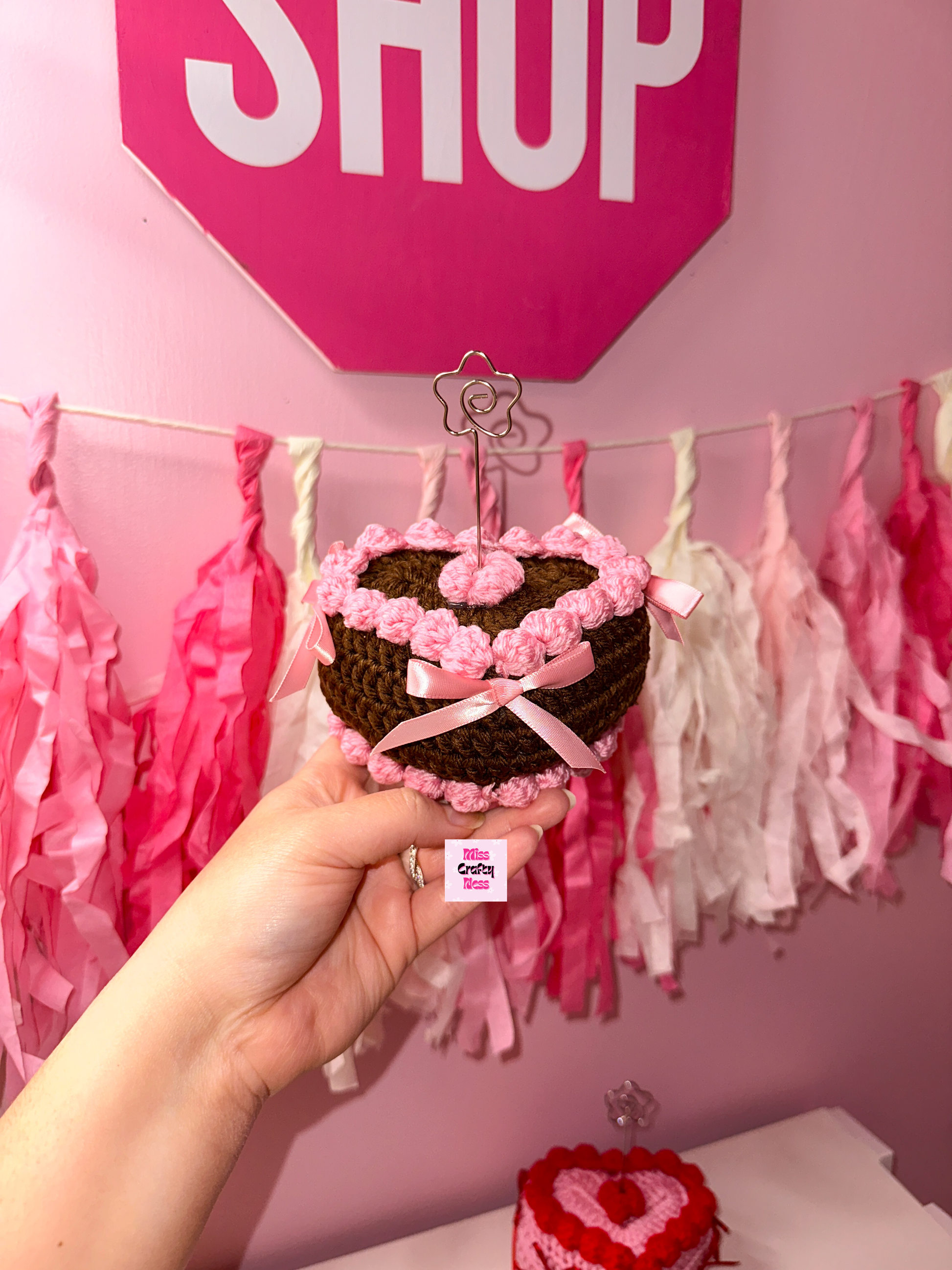 Brown Heart-shaped crocheted cake held in front of a pink 'SHOP' sign and tassel decorations.