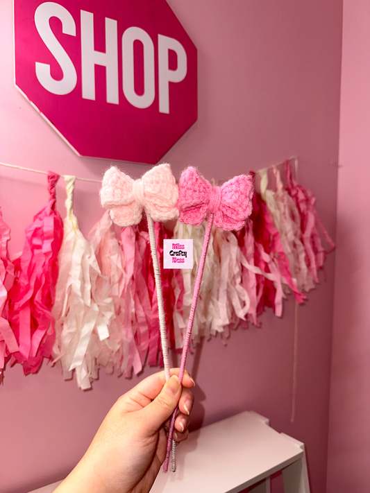 Pink bows held by a hand in front of a pink wall with 'SHOP' sign and tassel decorations.