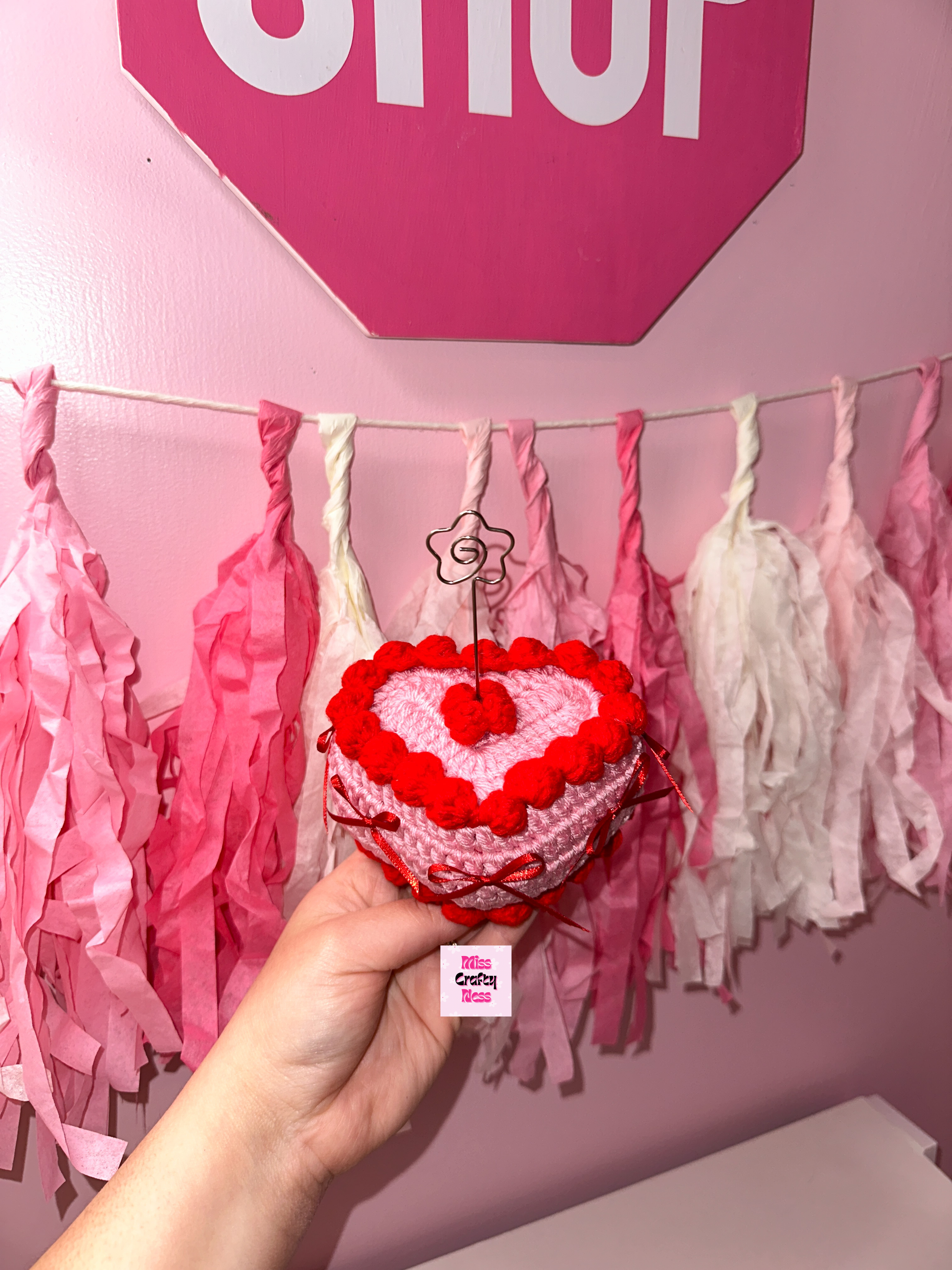 Pink Heart-shaped crocheted cake with red frosting held in front of a pink wall with tassels and a 'STOP' sign.