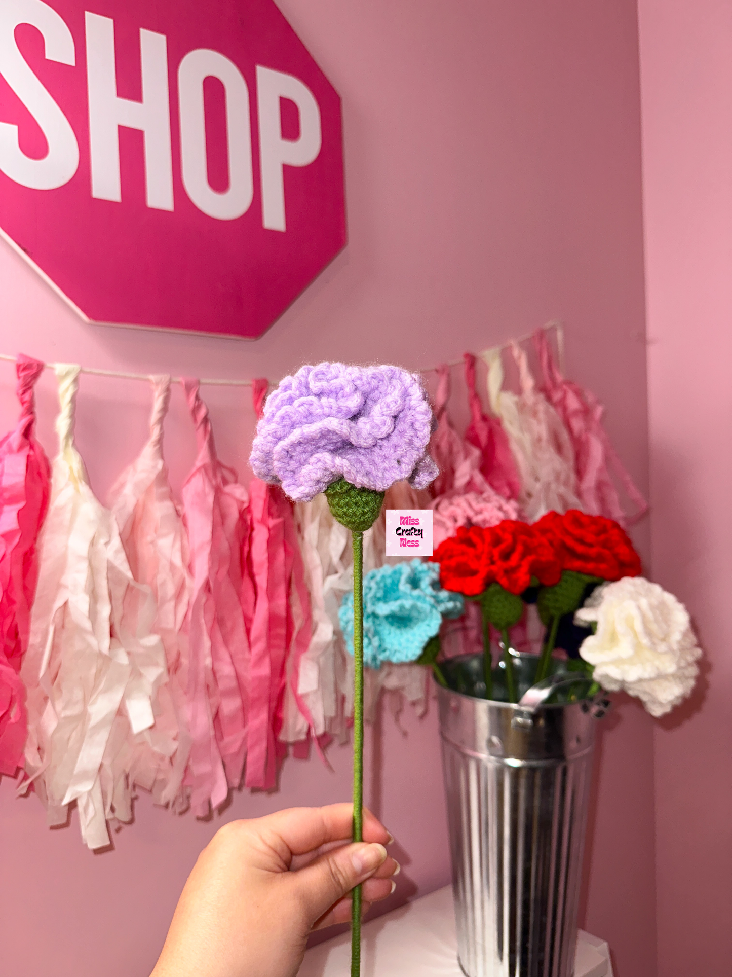 Hand holding a purple crocheted flower in front of a pink wall with tassels.