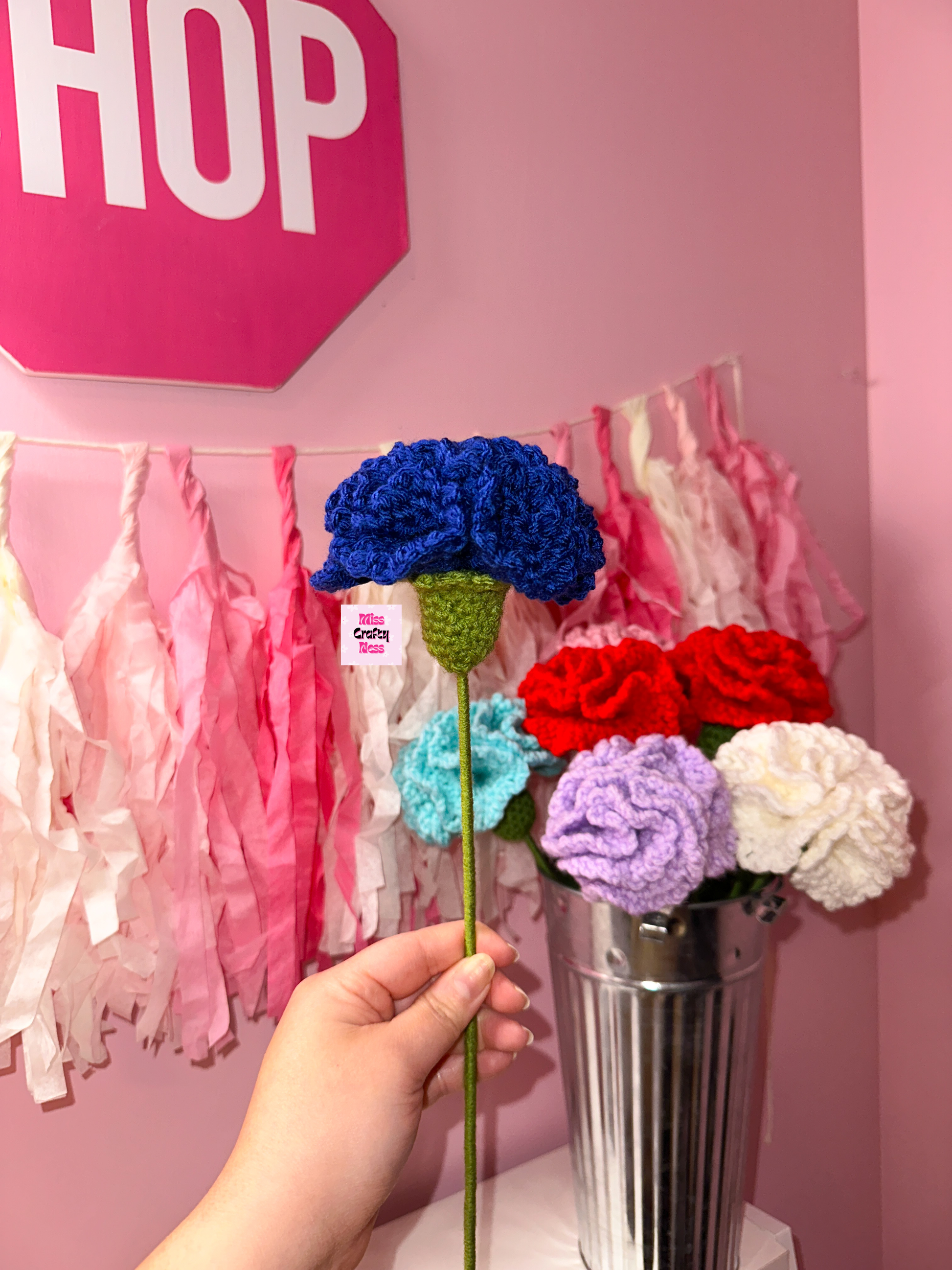 Blue crocheted flower held by a hand against a pink wall with 'SHOP' sign.