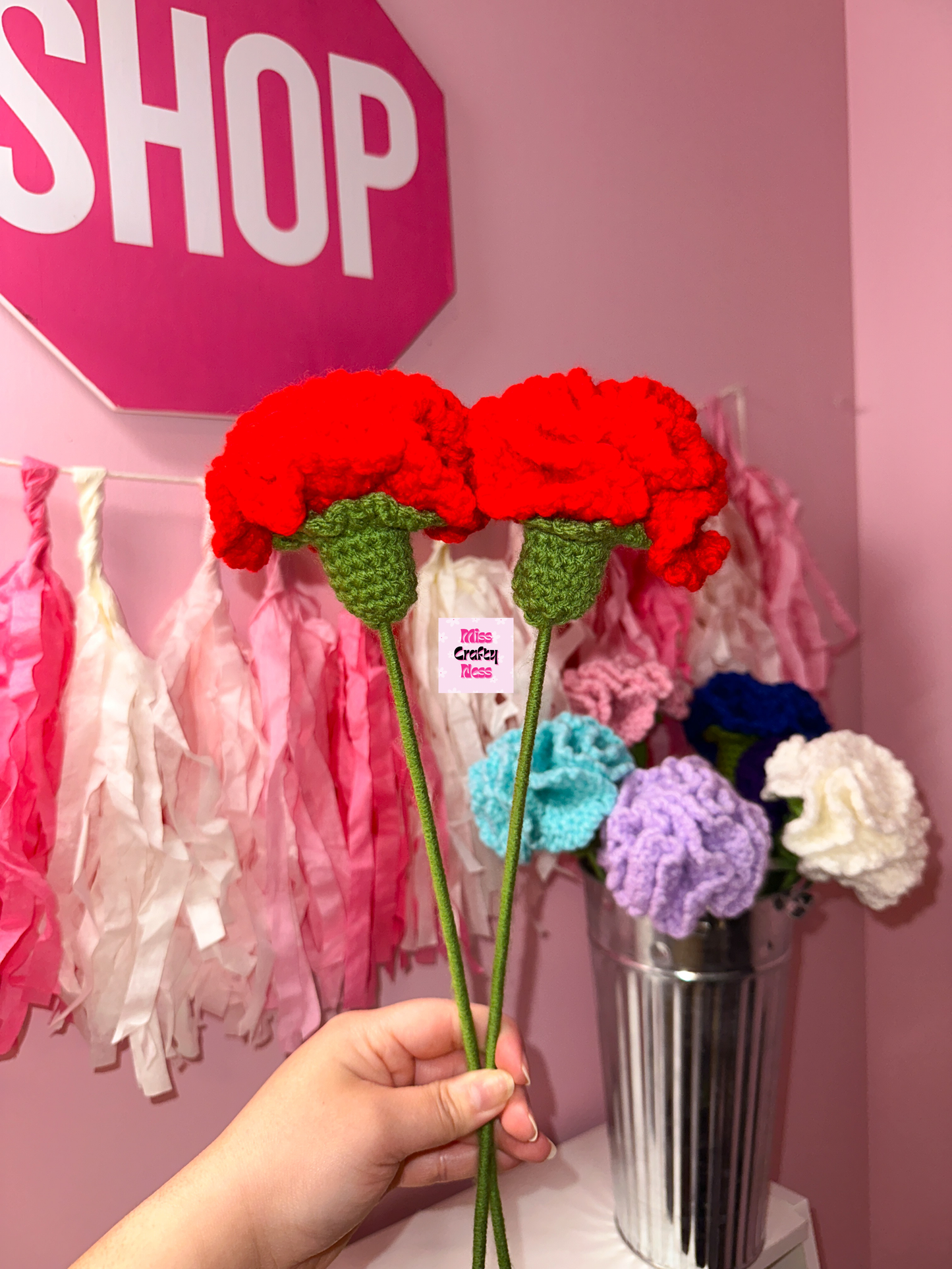 Red crochet flowers held by a hand with a pink 'SHOP' sign and colorful flowers in the background.