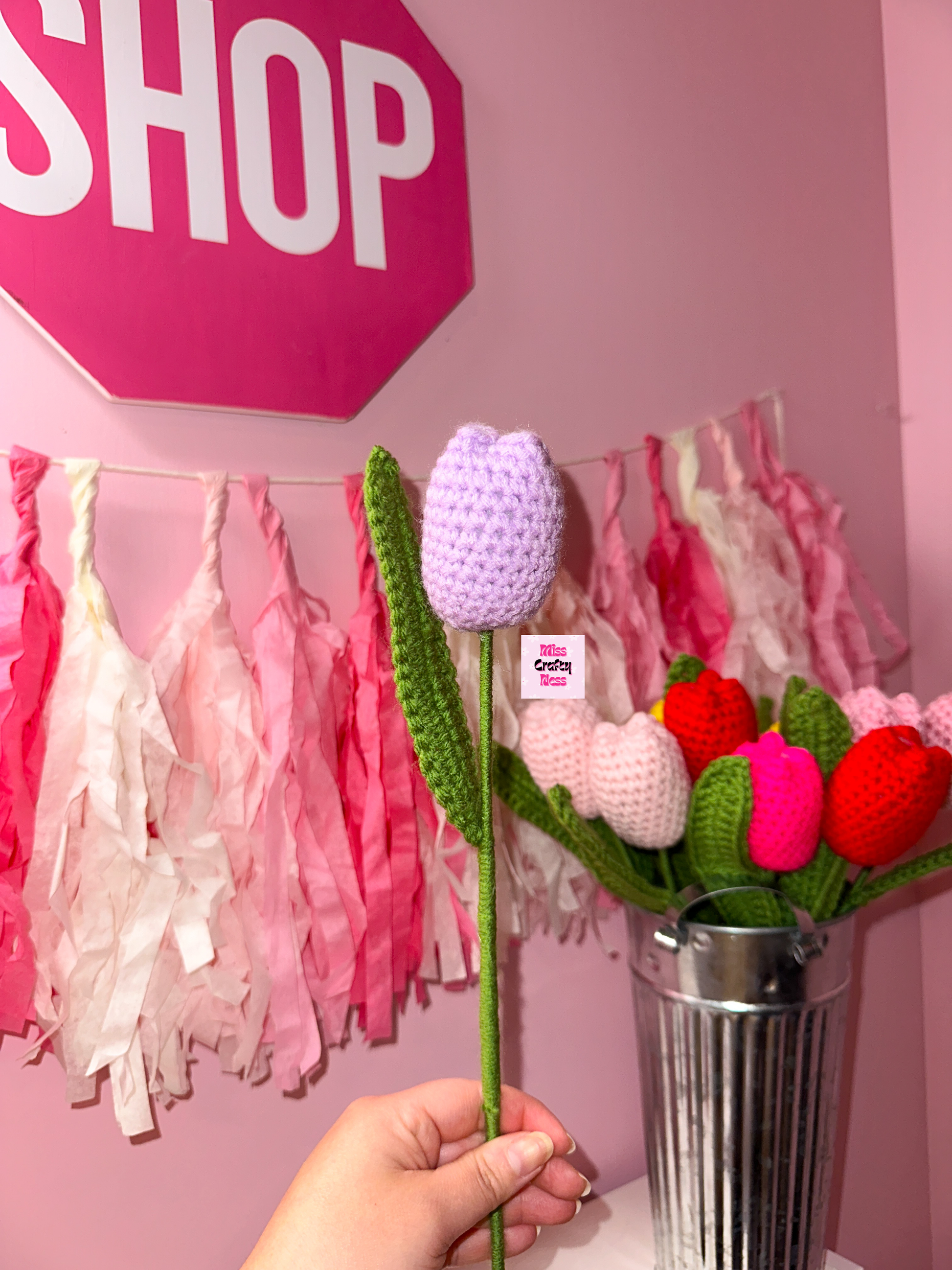 Hand holding a crocheted tulip flower with a pink 'SHOP' sign and decorative tassels in the background.