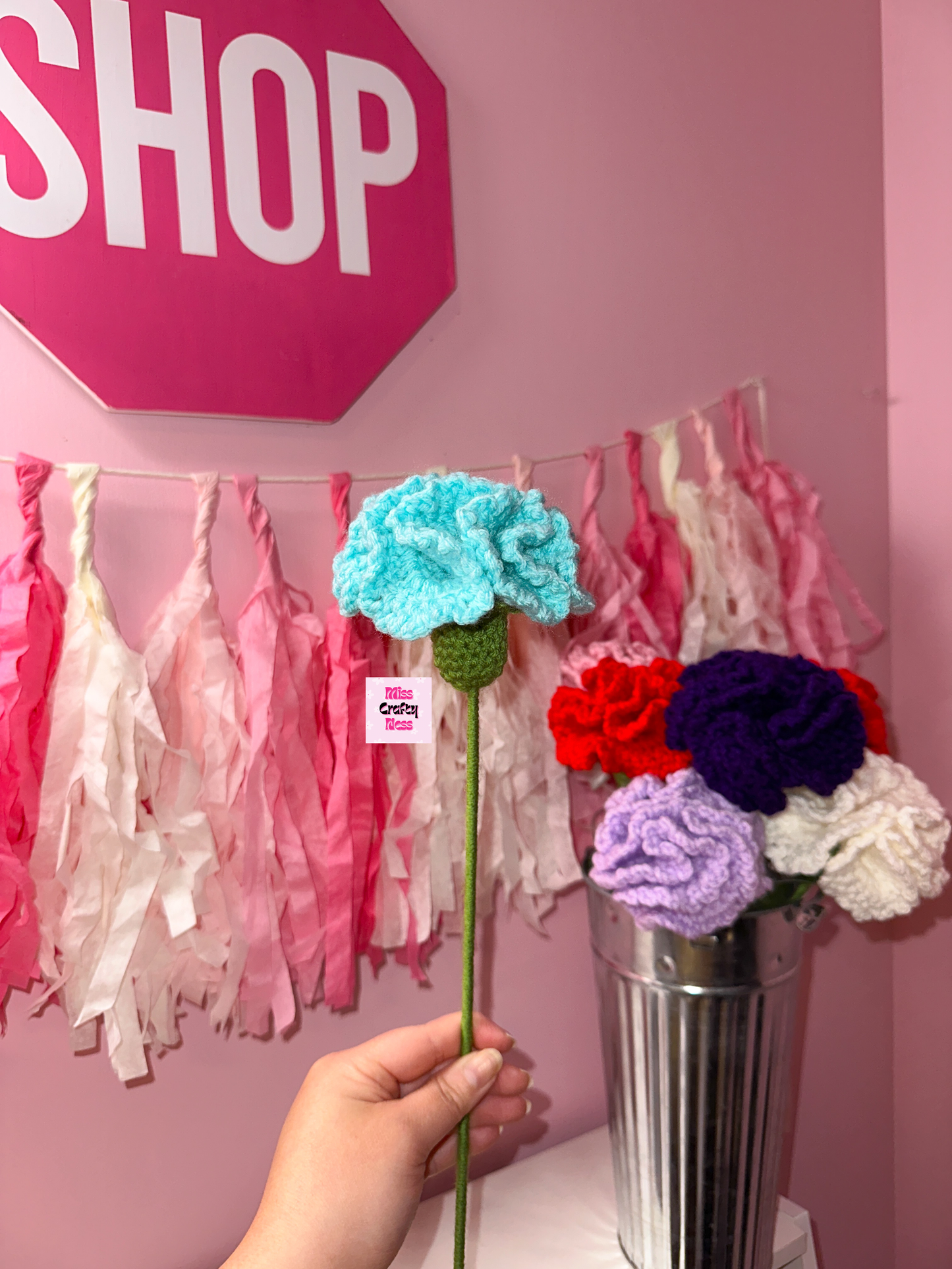 Hand holding a blue crocheted flower in front of a pink wall with tassels.