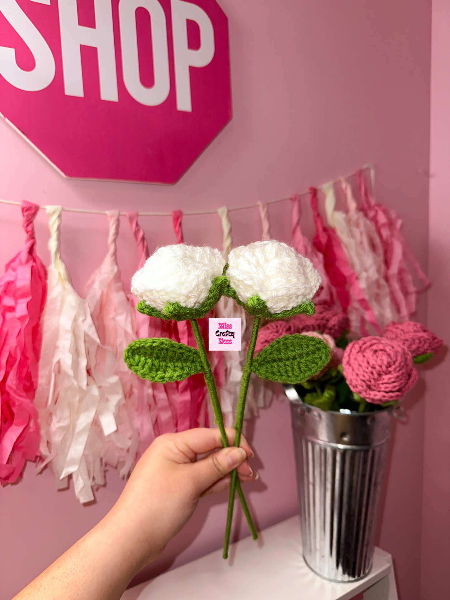 Hand holding a white crochet rose flower with a pink 'SHOP' sign and floral decorations in the background.