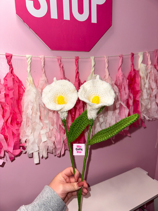 Hand holding two crocheted calla lily flowers with a pink 'SHOP' sign and decorative tassels in the background.