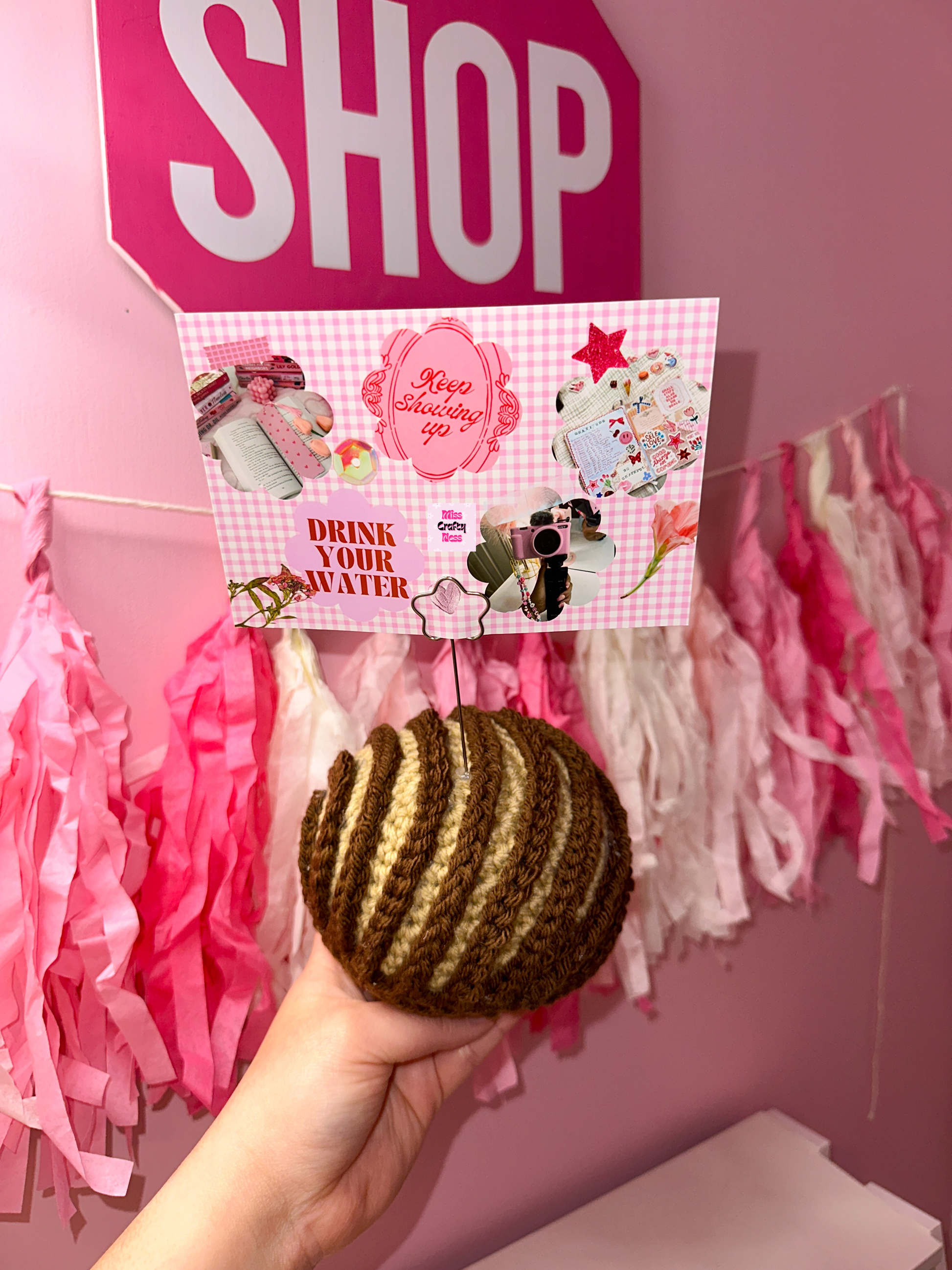 Crochet sweet bread photo holder item with pink and white tassels and a sign that reads ‘SHOP’ in the background.