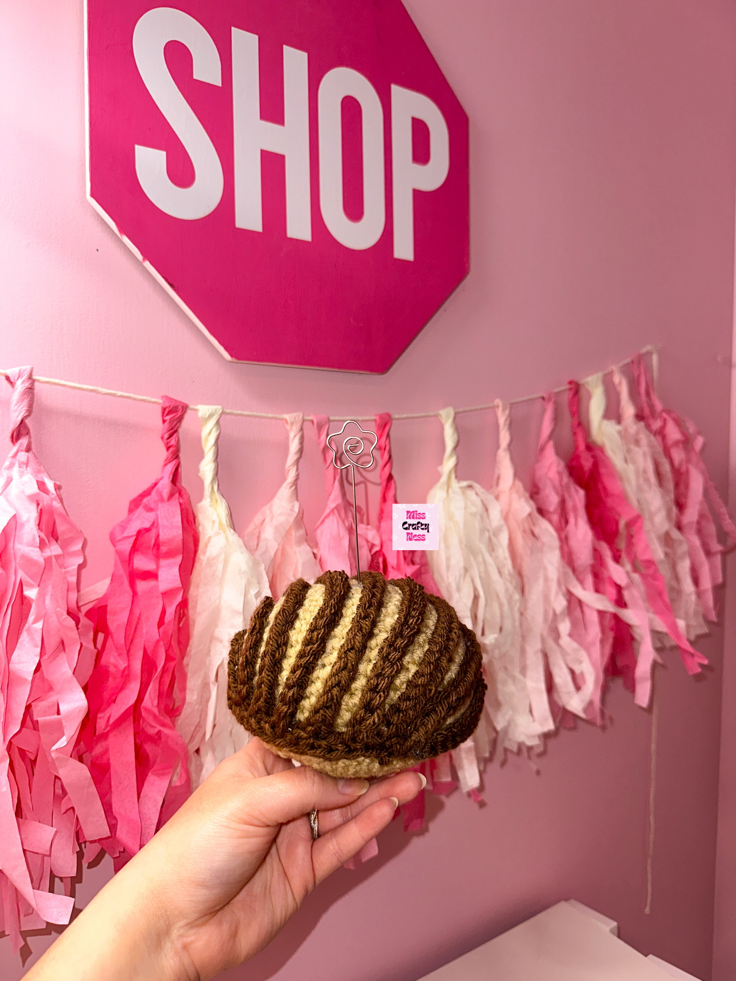 Crochet sweet bread photo holder item with pink and white tassels and a sign that reads ‘SHOP’ in the background.