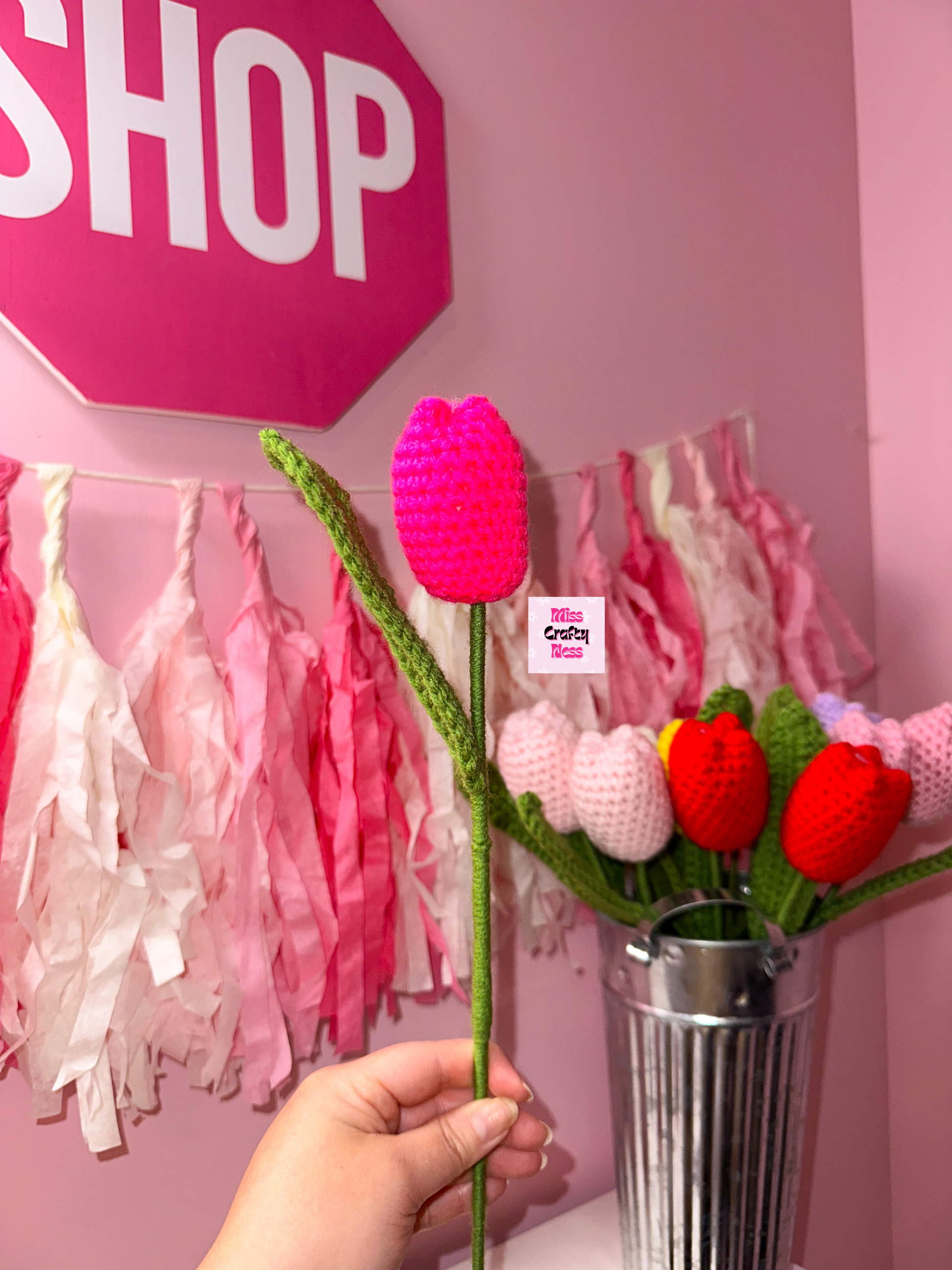 Hand holding a crocheted tulip flower with a pink 'SHOP' sign and decorative tassels in the background.