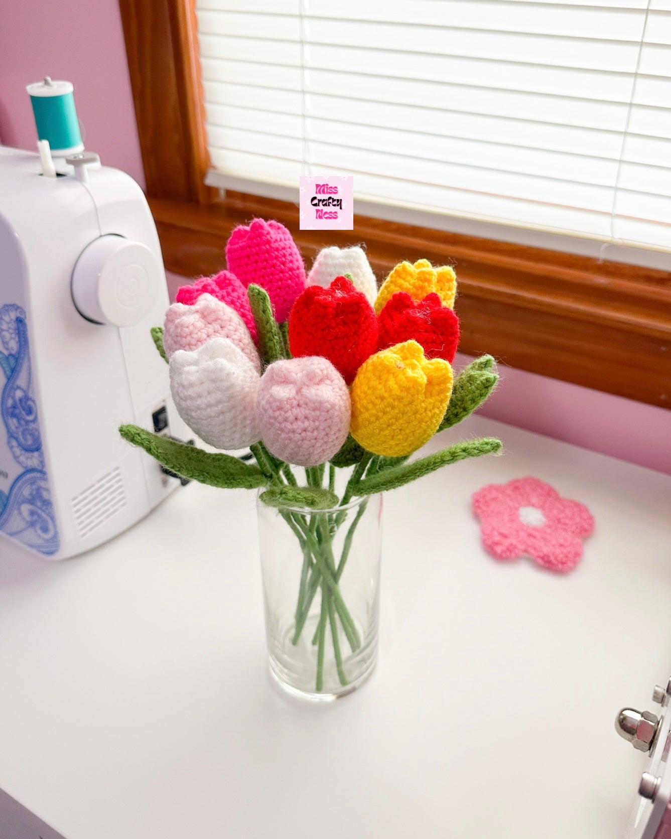 Colorful crocheted tulips in a vase by a sewing machine with a pink wall and window blinds in the background.