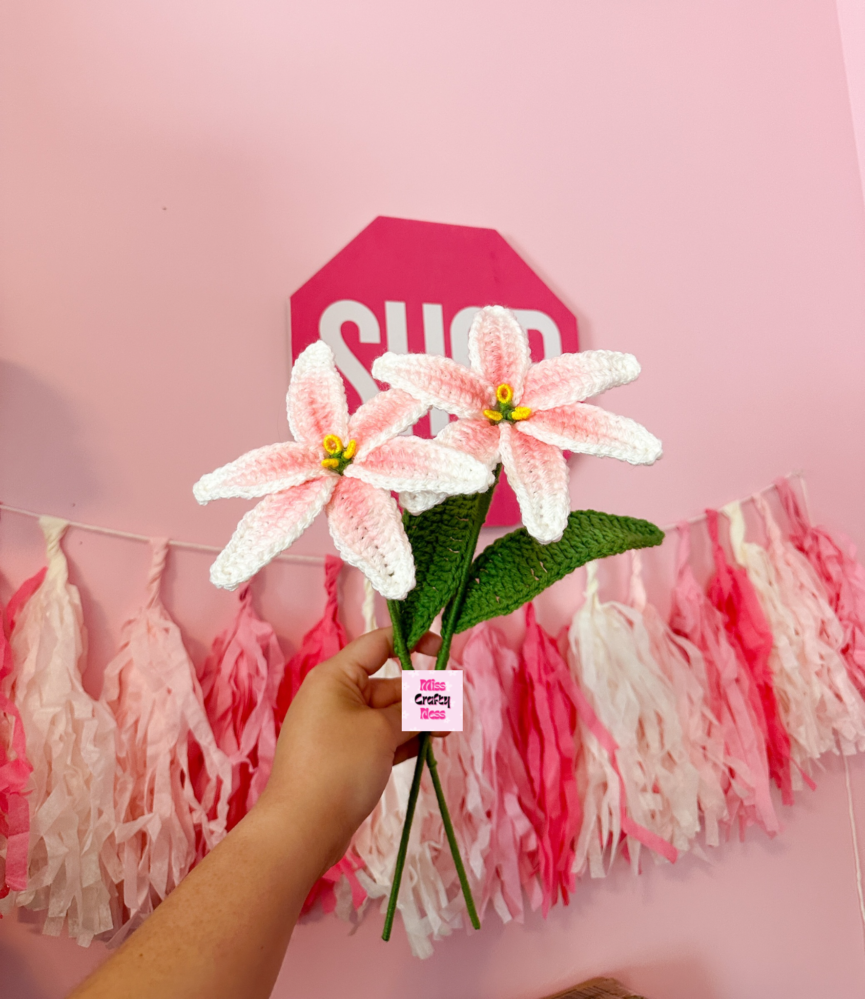 Hand holding two crochet Lily flowers against a pink wall with tassel decorations.