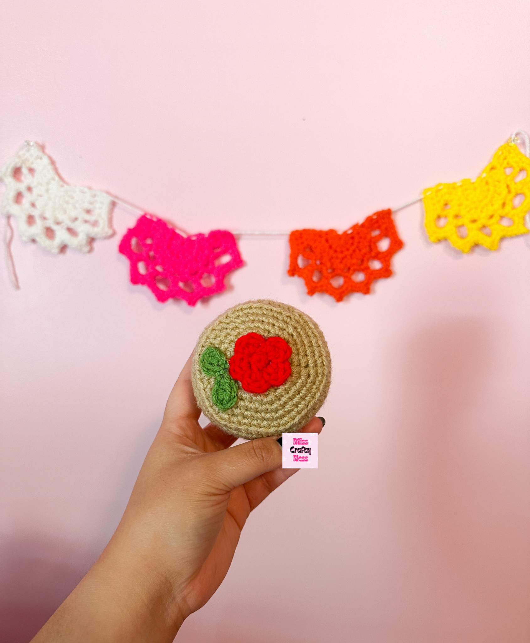Hand holding a crocheted round candy item with a red flower design against a pink background with colorful doily decoration
