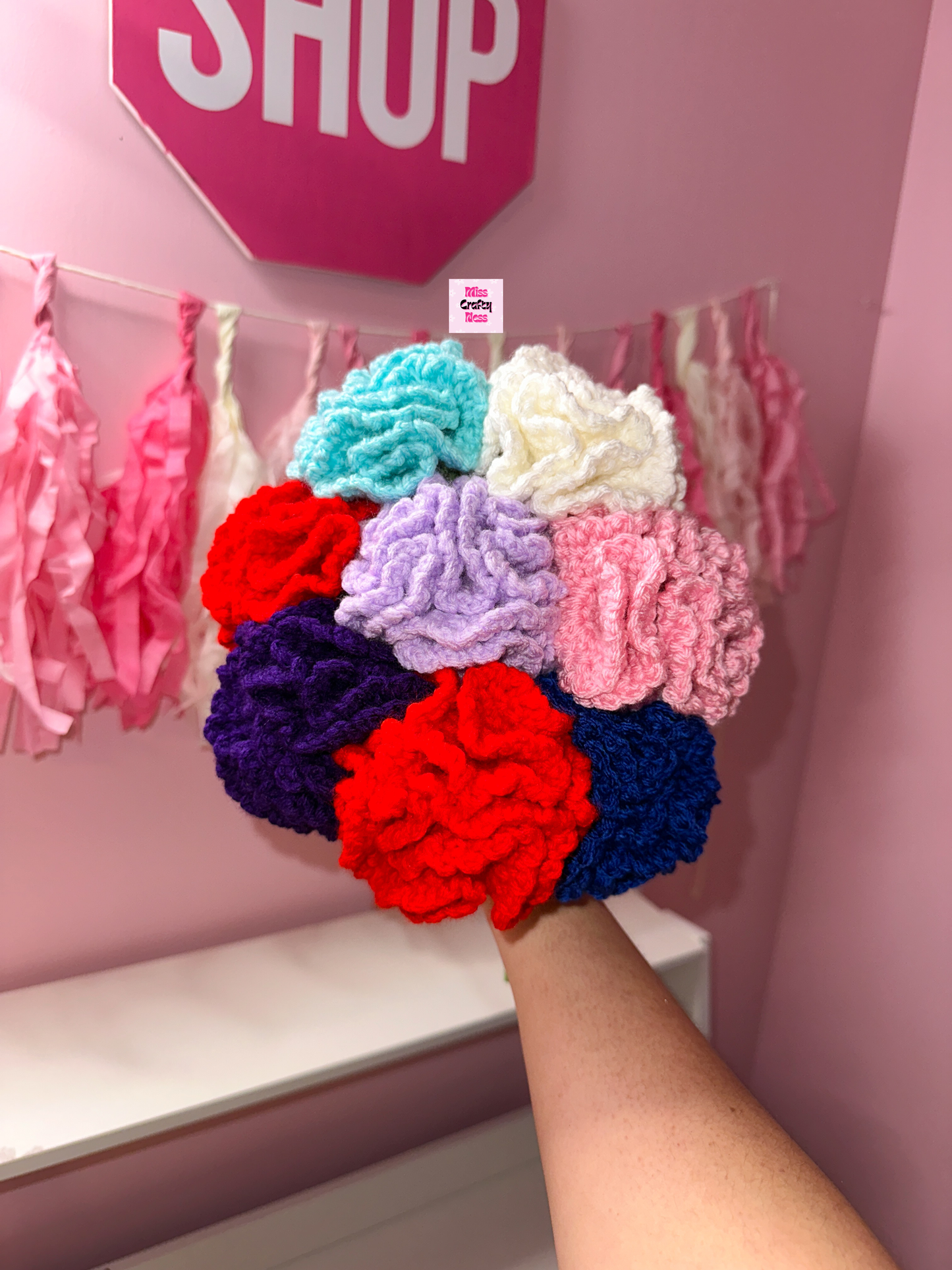 Colorful crocheted flowers held by a hand with a pink 'SHOP' sign in the background.