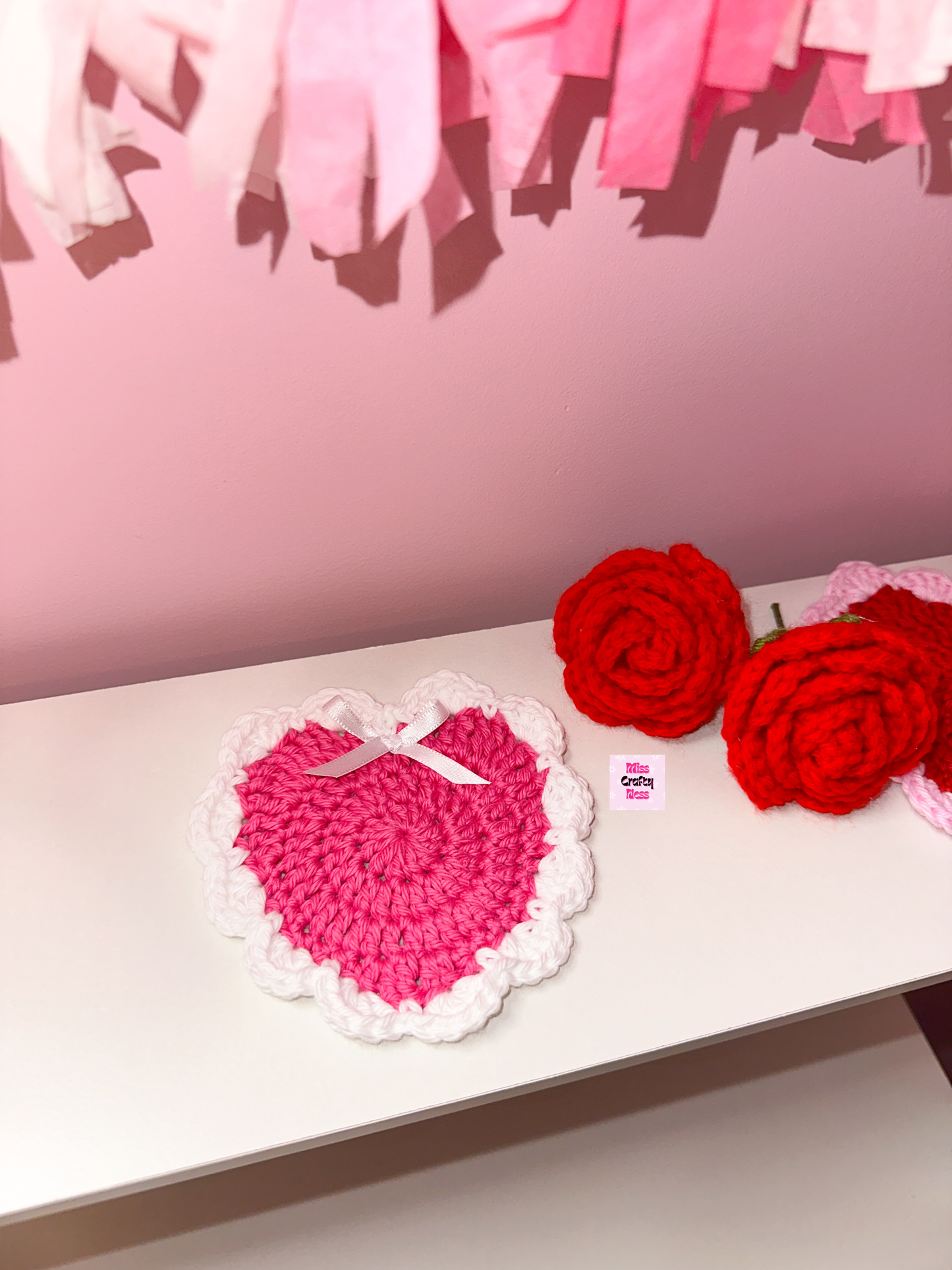 Heart-shaped pink crocheted item and red roses on a white surface with a pink background