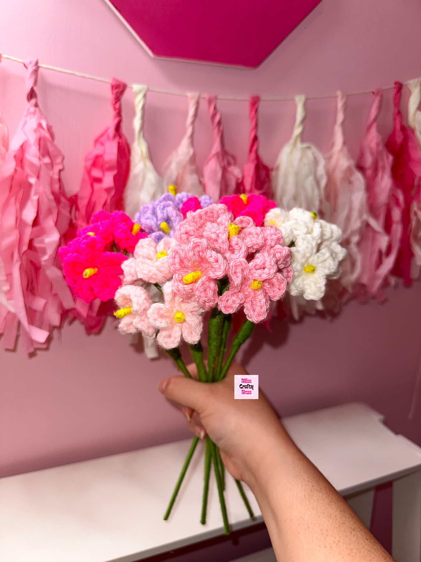 Bouquet of colorful crochet flowers held against a pink wall with tassels.